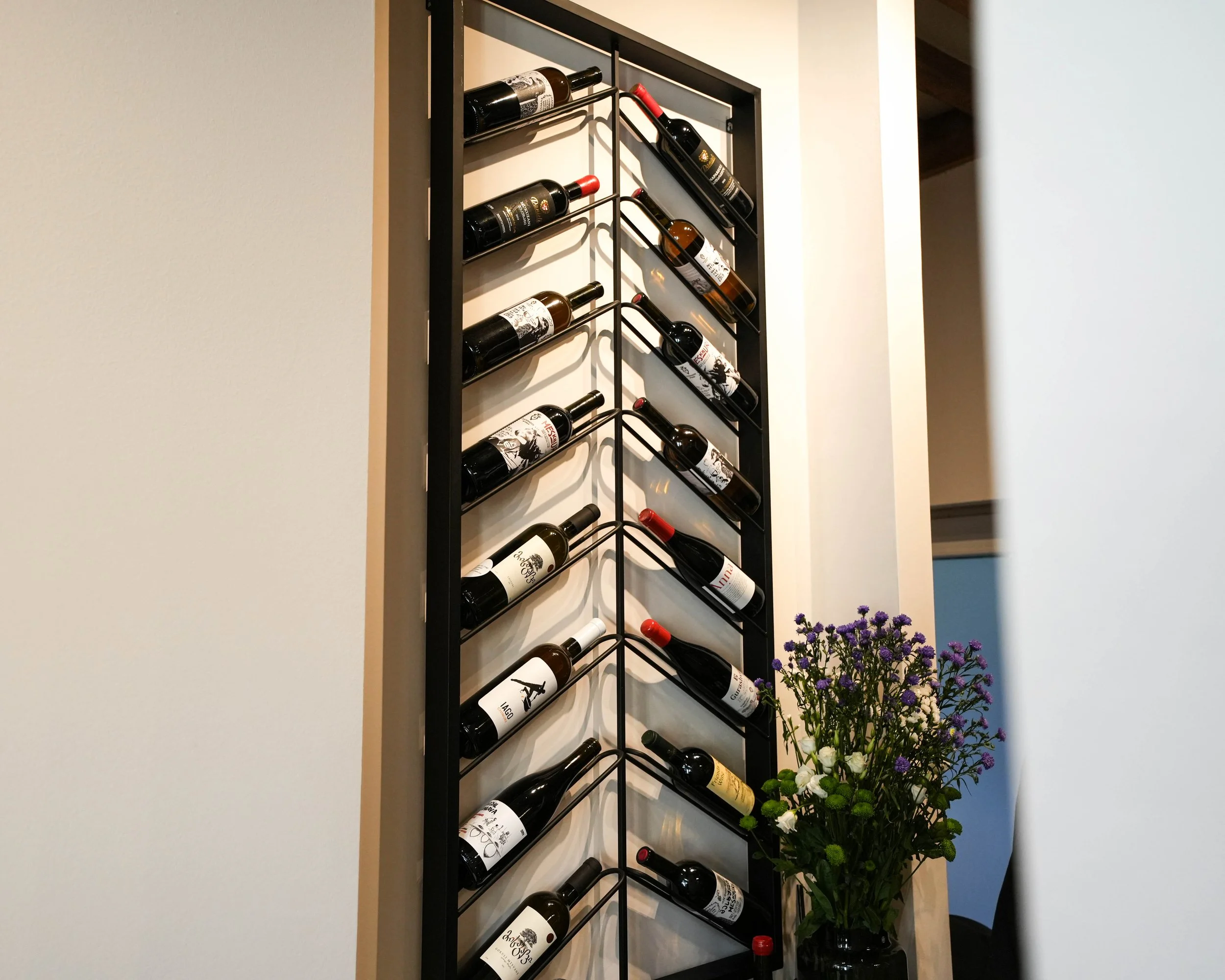 A vertical wine rack with multiple bottles of wine stored on black metal racks against a white wall, with purple and white flowers in a vase in the lower right corner.