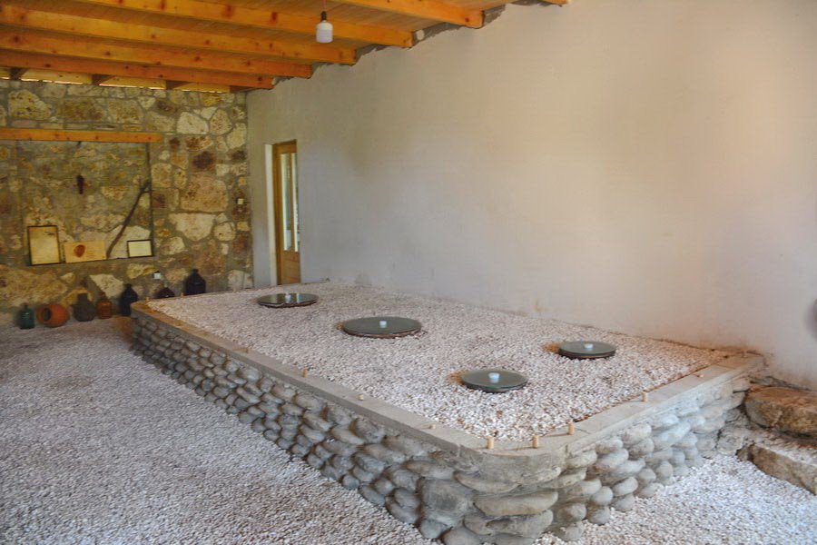 Interior of a rustic room with stone and plaster walls, a stone fireplace, and a wooden ceiling, featuring a bed with a stone border and several decorative pots on a shelf.