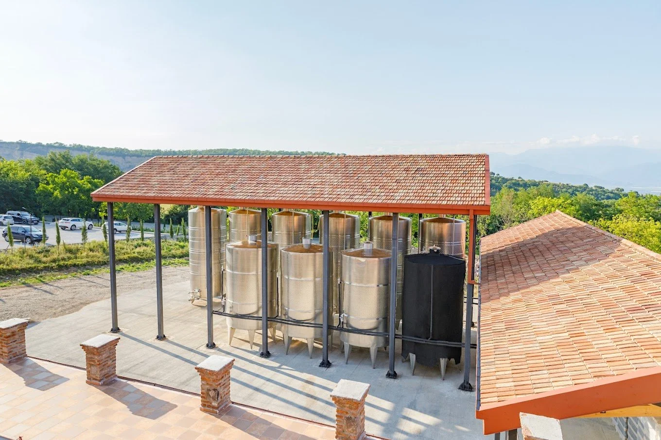 Multiple stainless steel tanks under a red-tile roof, part of a winery or brewery, with a gravel parking lot and green landscape in the background.