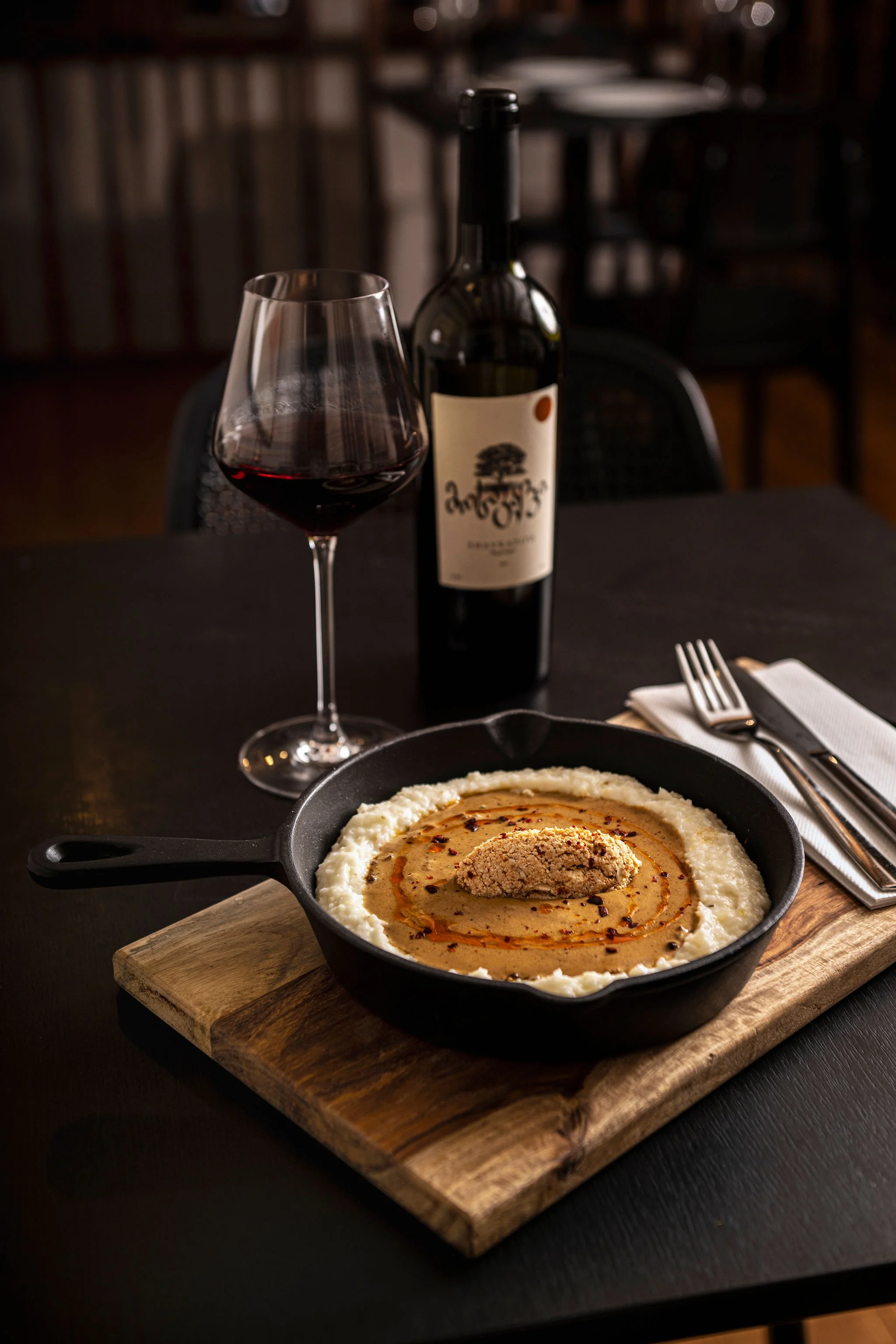 A glass of red wine and a bottle of wine on a table, with a dish of hummus topped with a scoop of hummus and paprika, all set on a wooden cutting board in a dimly lit restaurant.