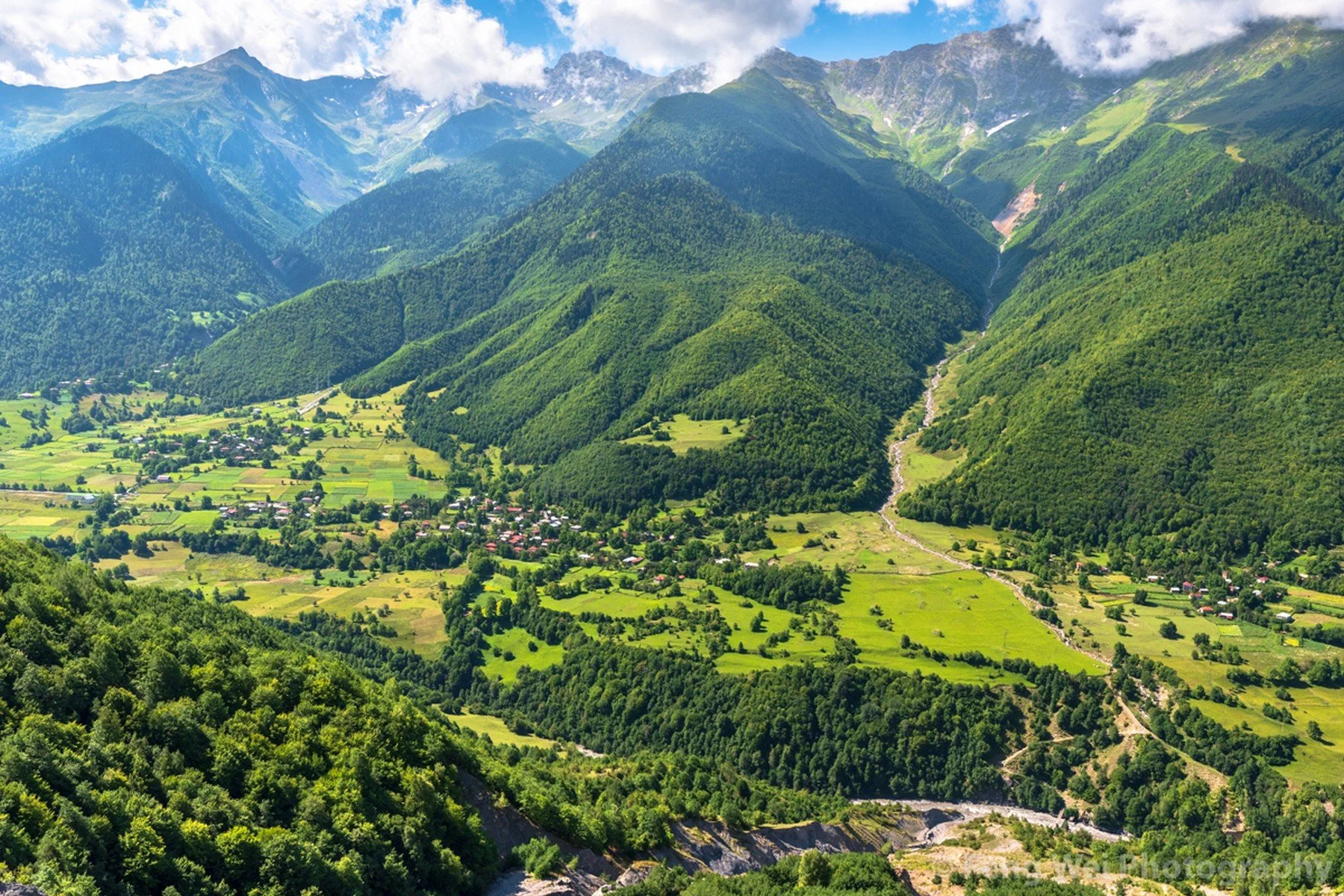 Scenic view of a lush green valley with small houses, surrounded by tall mountains with dense forests and a partly cloudy sky.