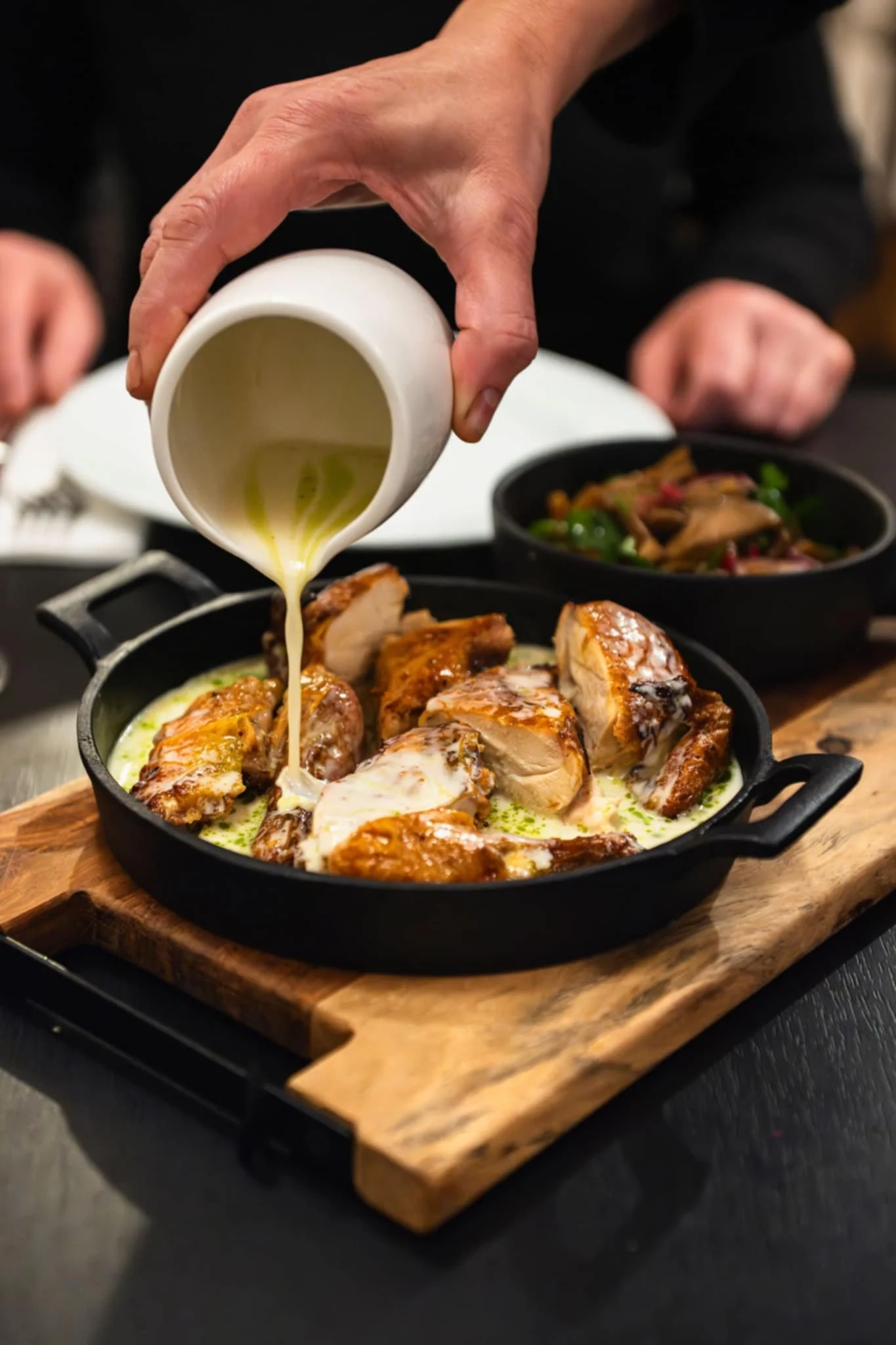 A person pouring a creamy sauce over roasted chicken pieces in a cast iron skillet on a wooden cutting board, with side dishes in black bowls in the background.