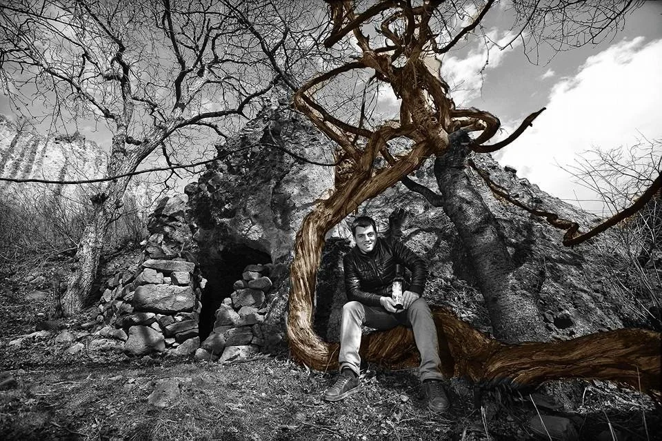 A man sitting on a fallen tree trunk in front of a rocky hillside and a cave, with leafless trees and branches around, in black and white.