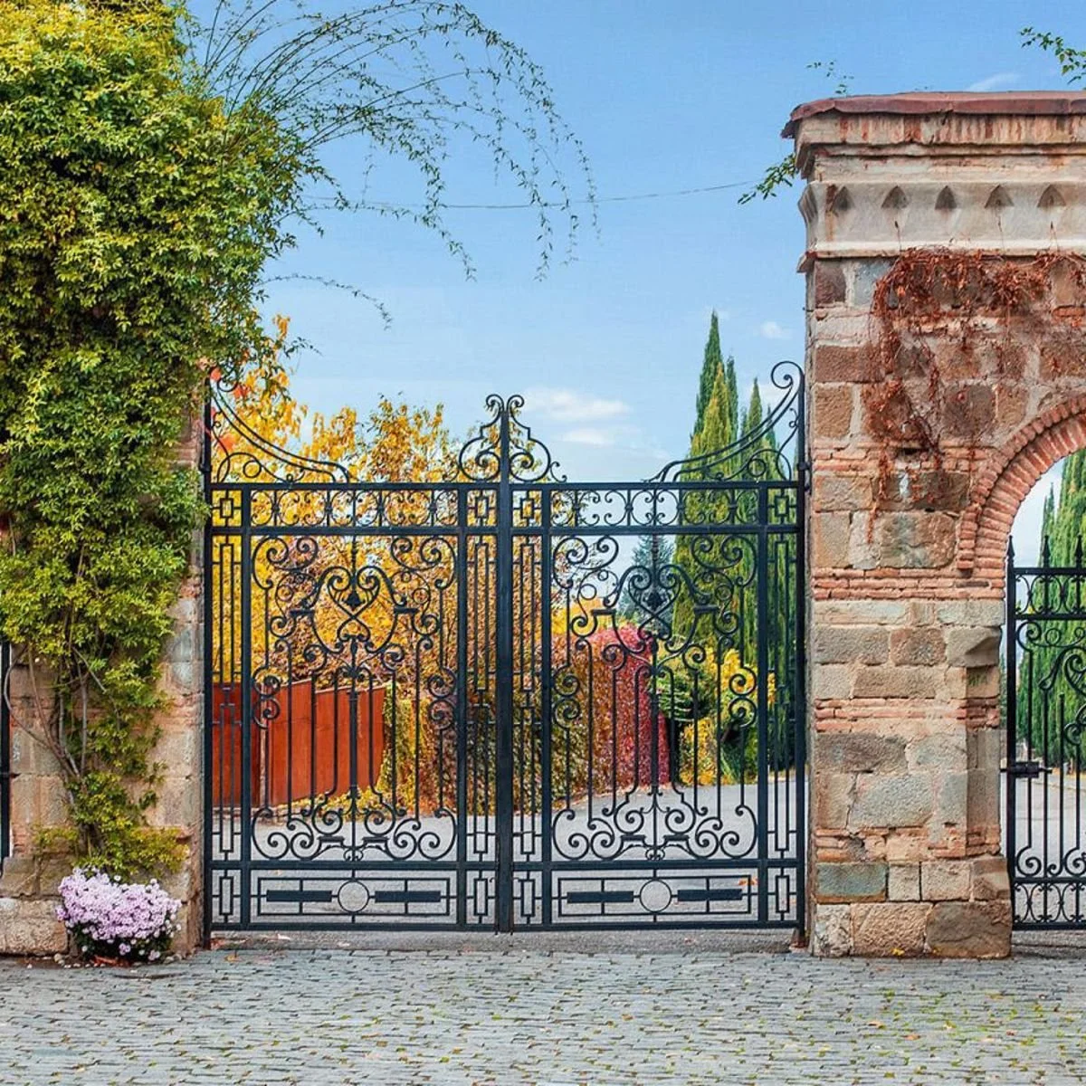 Black wrought iron gate with ornate scrollwork between two stone pillars, with trees and a street behind.