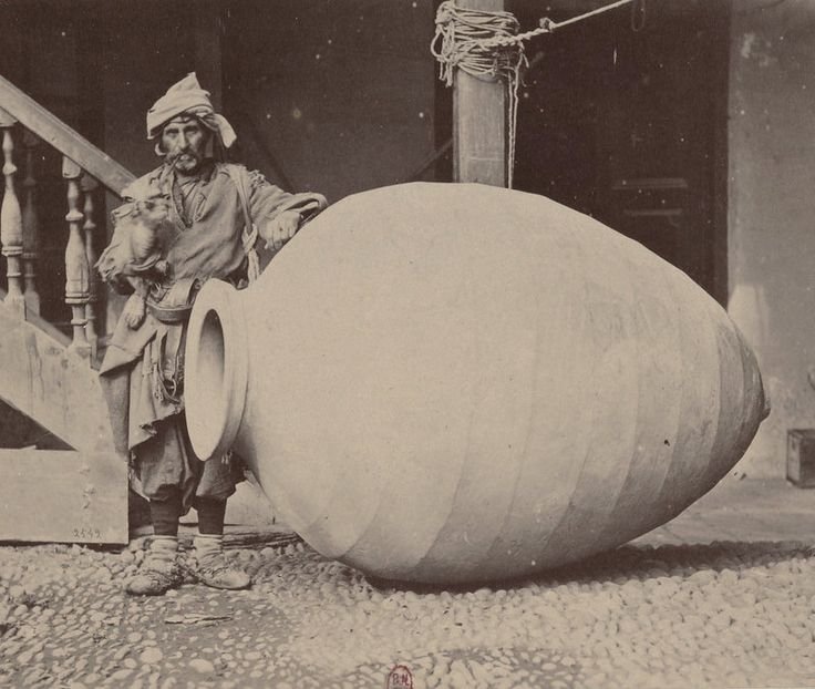 A man in traditional attire standing next to an oversized, partially carved clay or ceramic vessel indoors, with a dog resting its head on his shoulder.