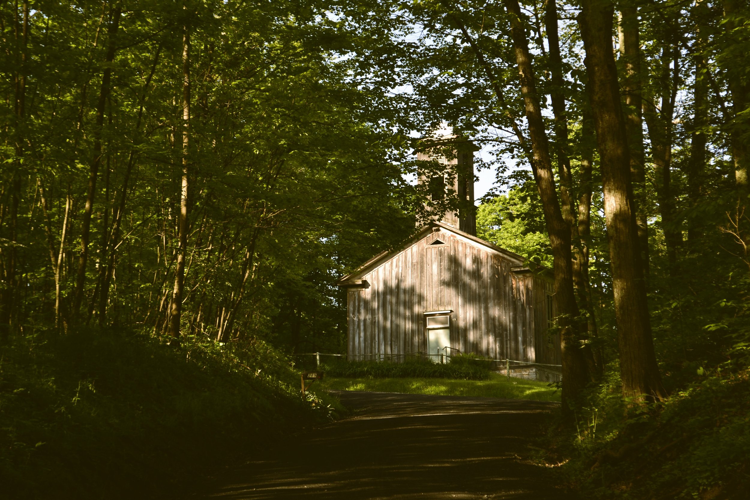 A small wooden church at the end of a woodland road,  with sunlight filtering through the leaves.
