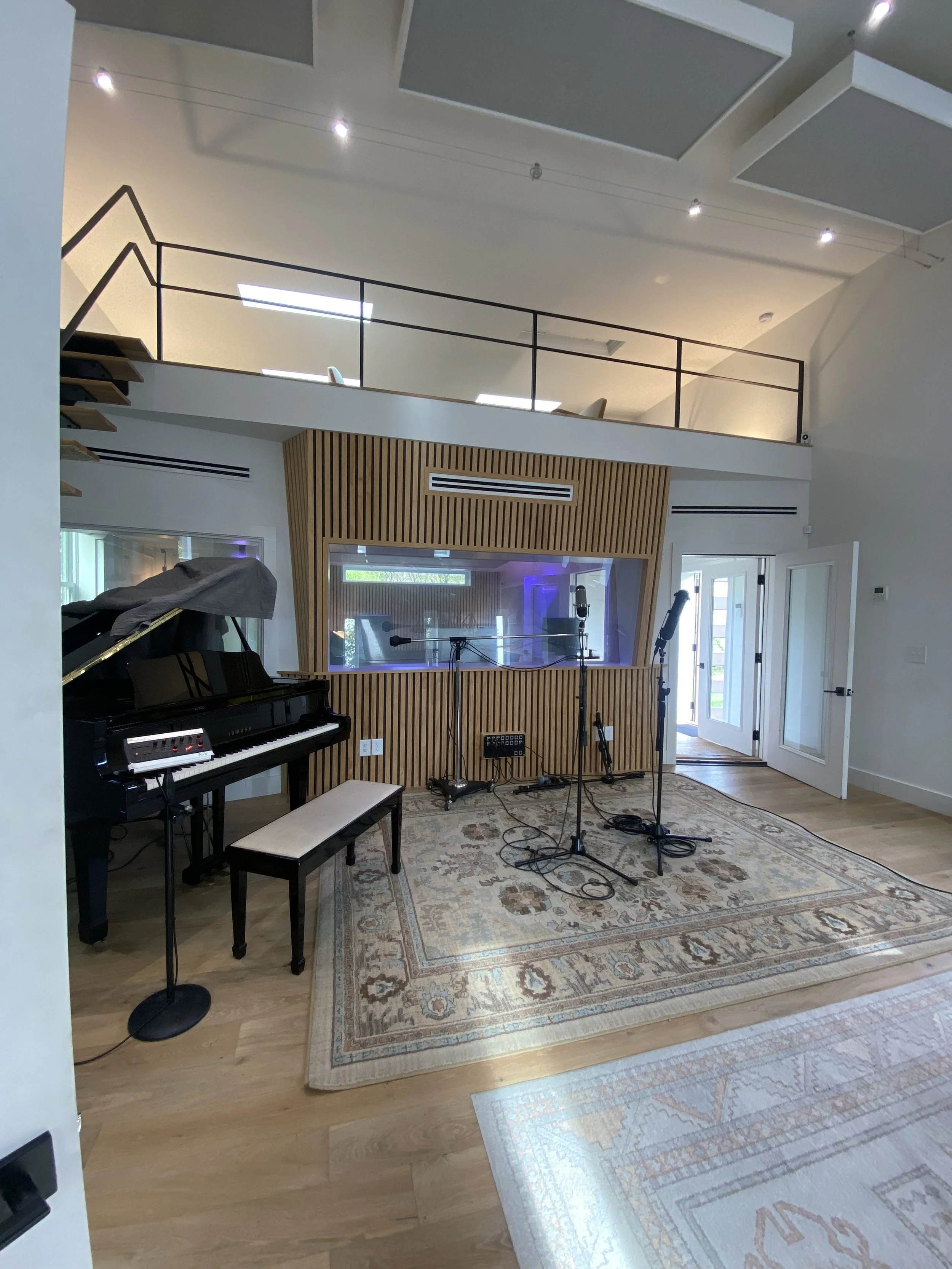 A music recording studio with a grand piano, microphone stands, and musical equipment set up for recording, with a wooden accent wall, a patterned rug, and a door leading outside.