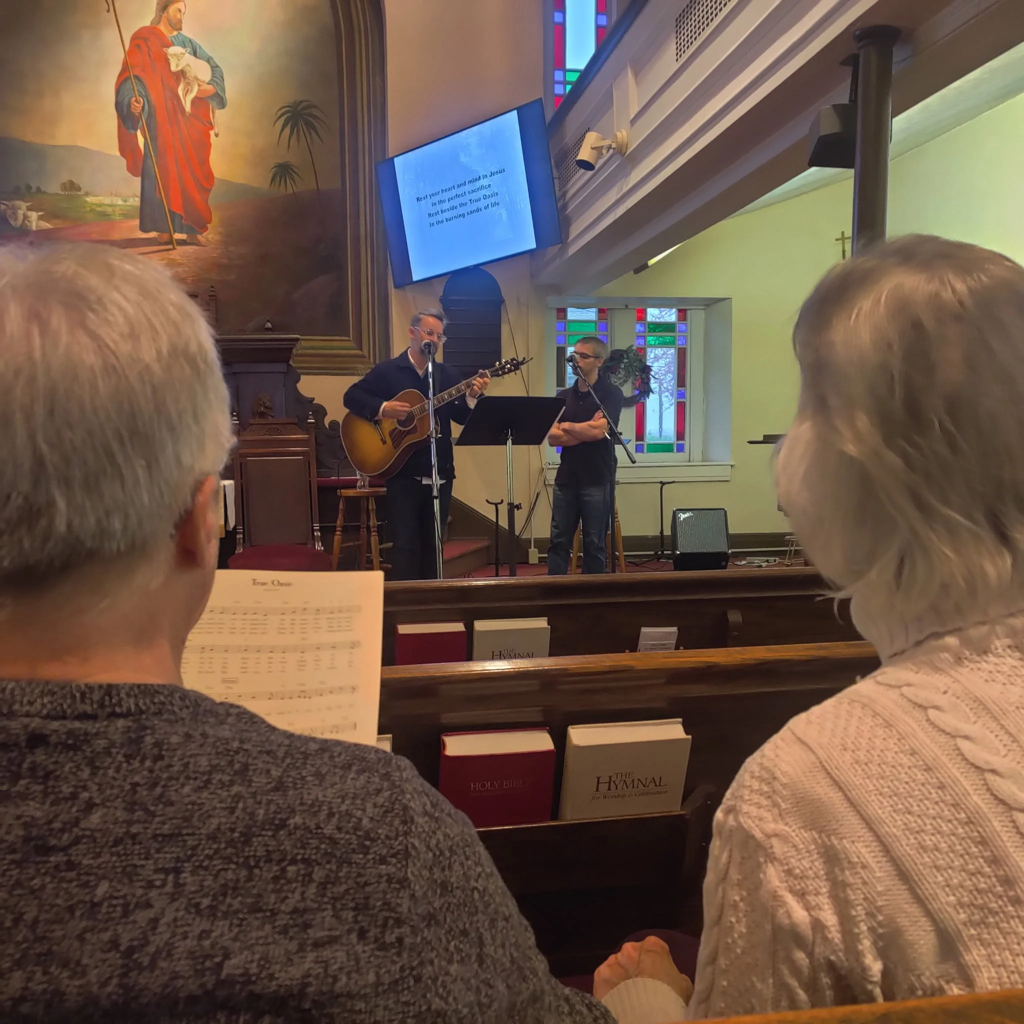 People in a church singing and playing guitar during a service. Visible stained glass windows and religious paintings decorate the space.