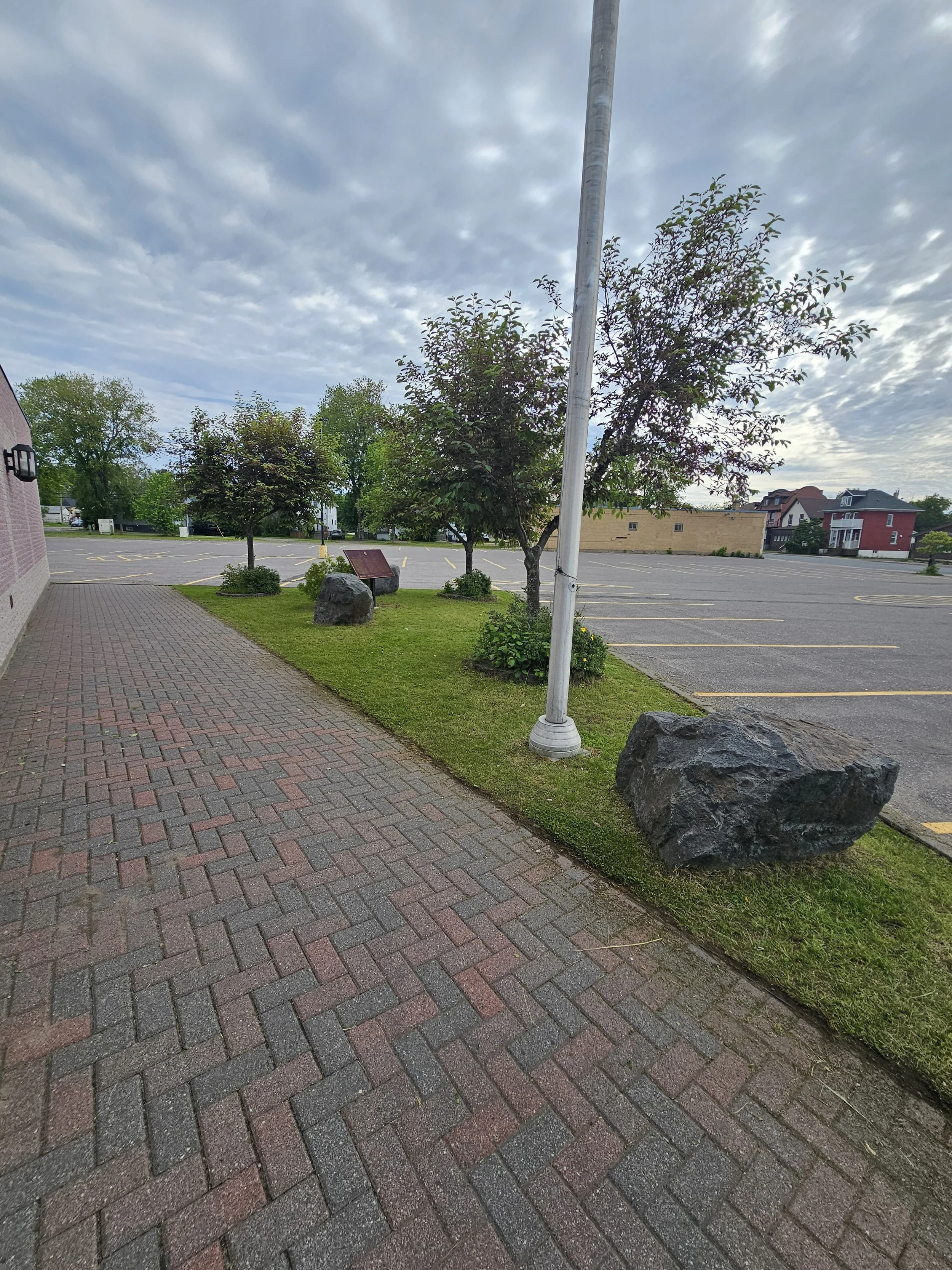 A parking lot with a brick sidewalk, three trees, large rocks, a pole, and a row of houses in the background under a cloudy sky.