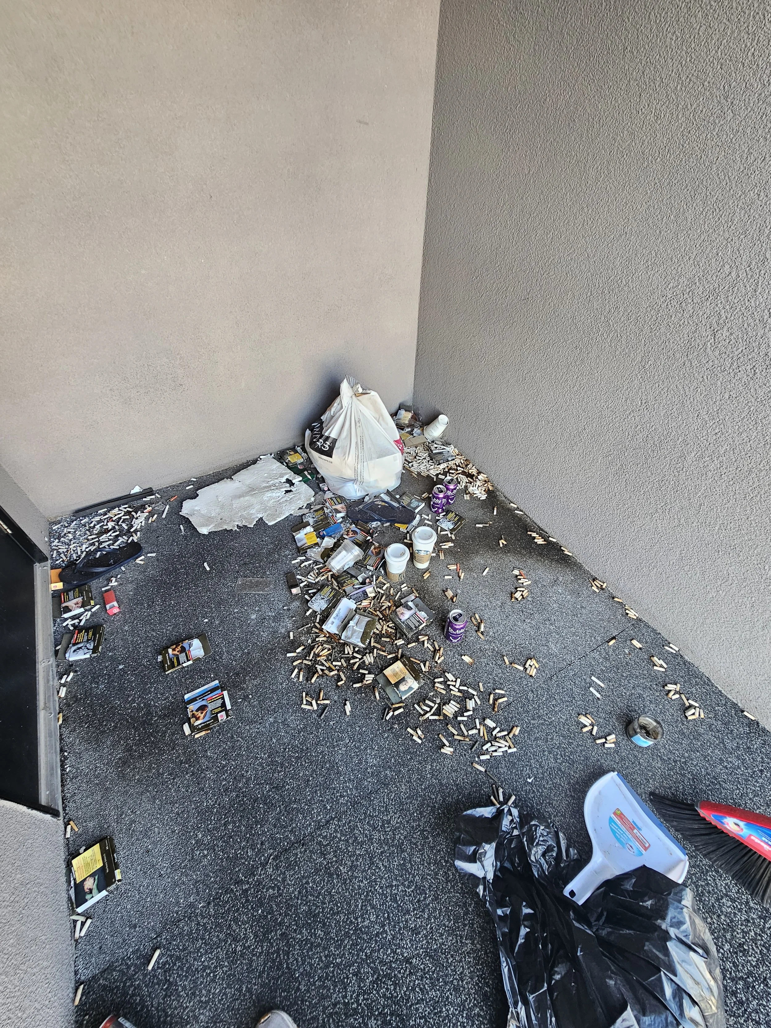 An overfilled trash area with numerous cigarette packs, cigarette butts, and empty coffee cups scattered on the ground, in a corner of a room.