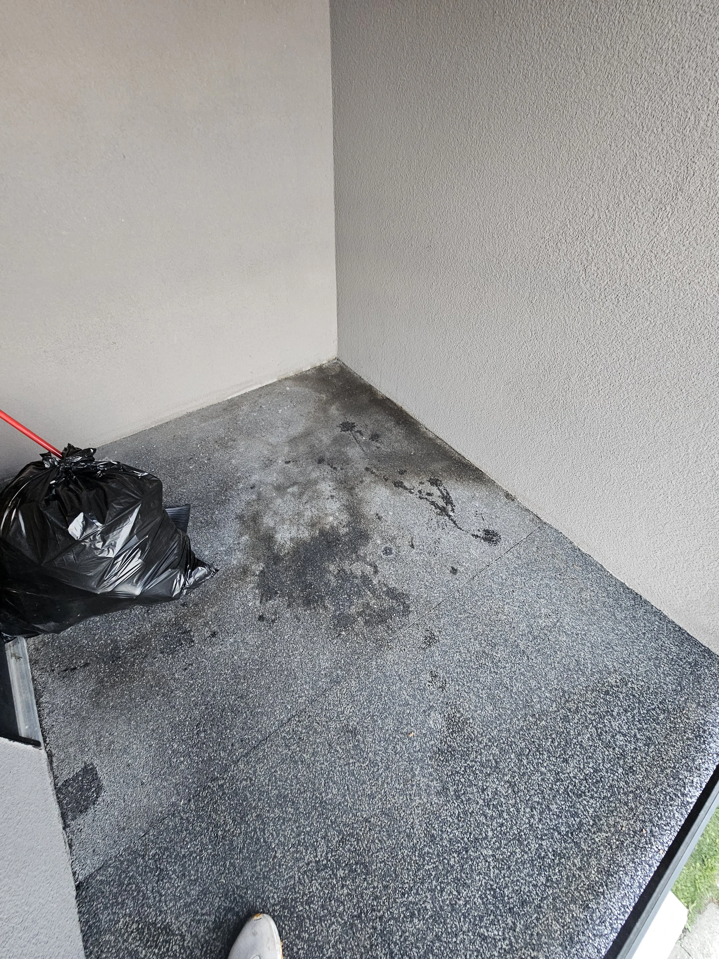 A corner of a room with beige walls, a rough-textured floor, and a black trash bag on the left side.