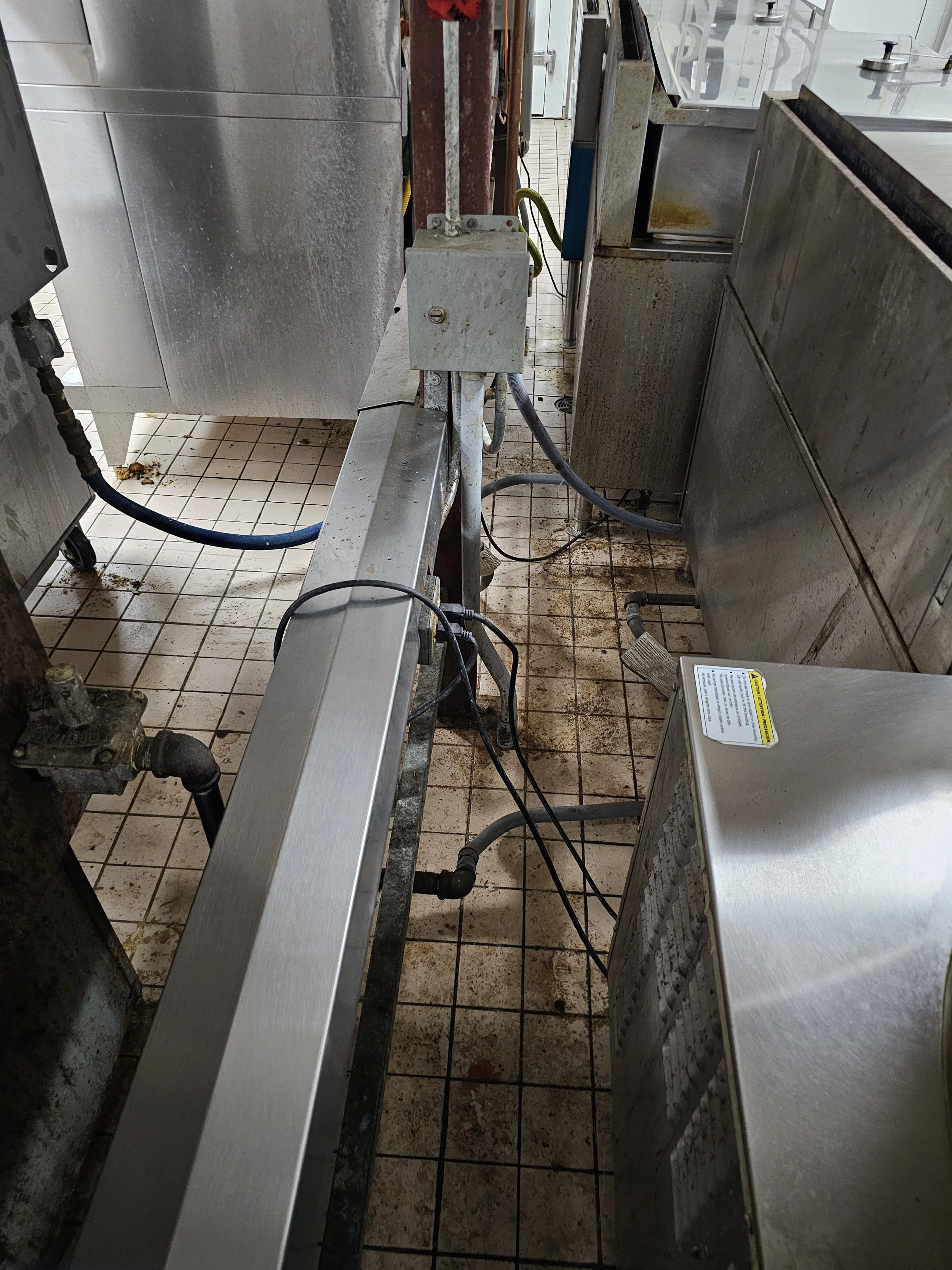 A commercial kitchen with stainless steel countertops, pipes, and a tiled floor that is dirty with spots and spills. There are cables and a small appliance with a warning label on it.