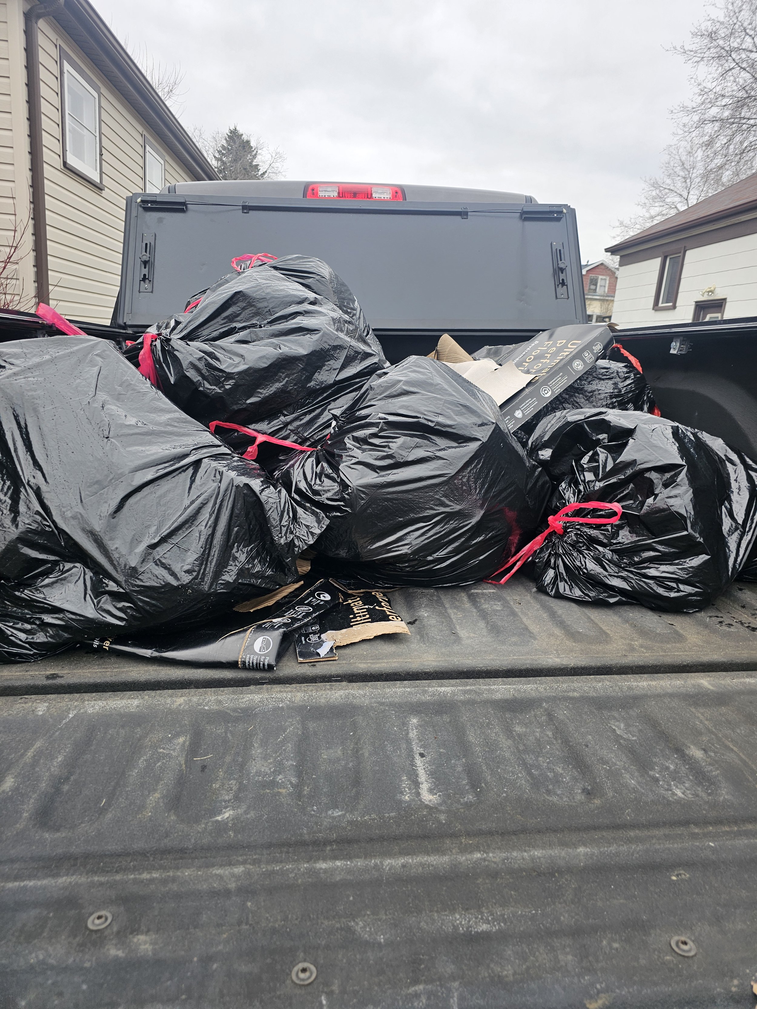 The back of a truck bed filled with black trash bags tied with pink ties and some cardboard boxes, with neighboring houses visible in the background under an overcast sky.