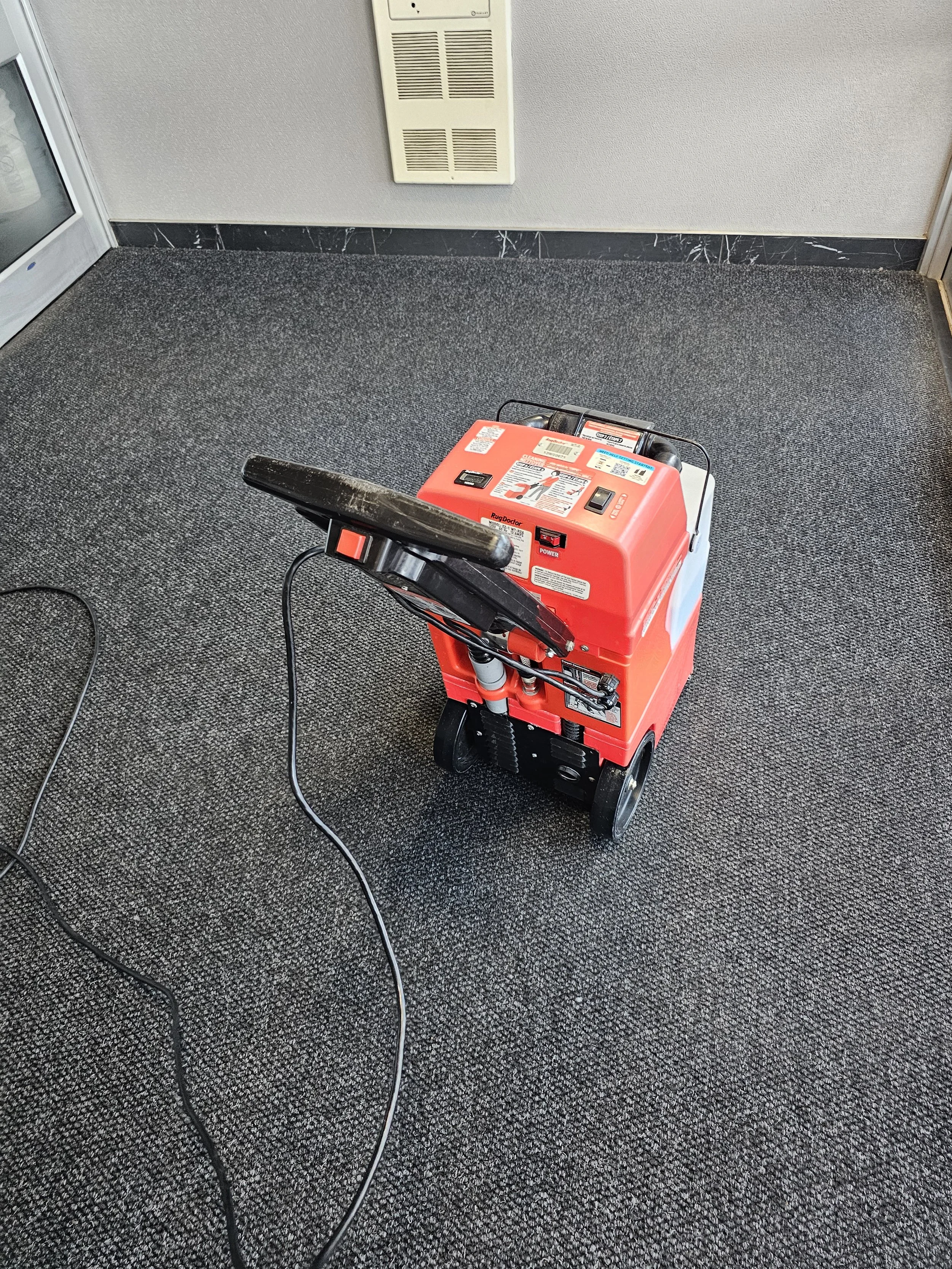 An orange portable carpet cleaning machine with black handles on a gray carpeted floor, near a wall with a vent.