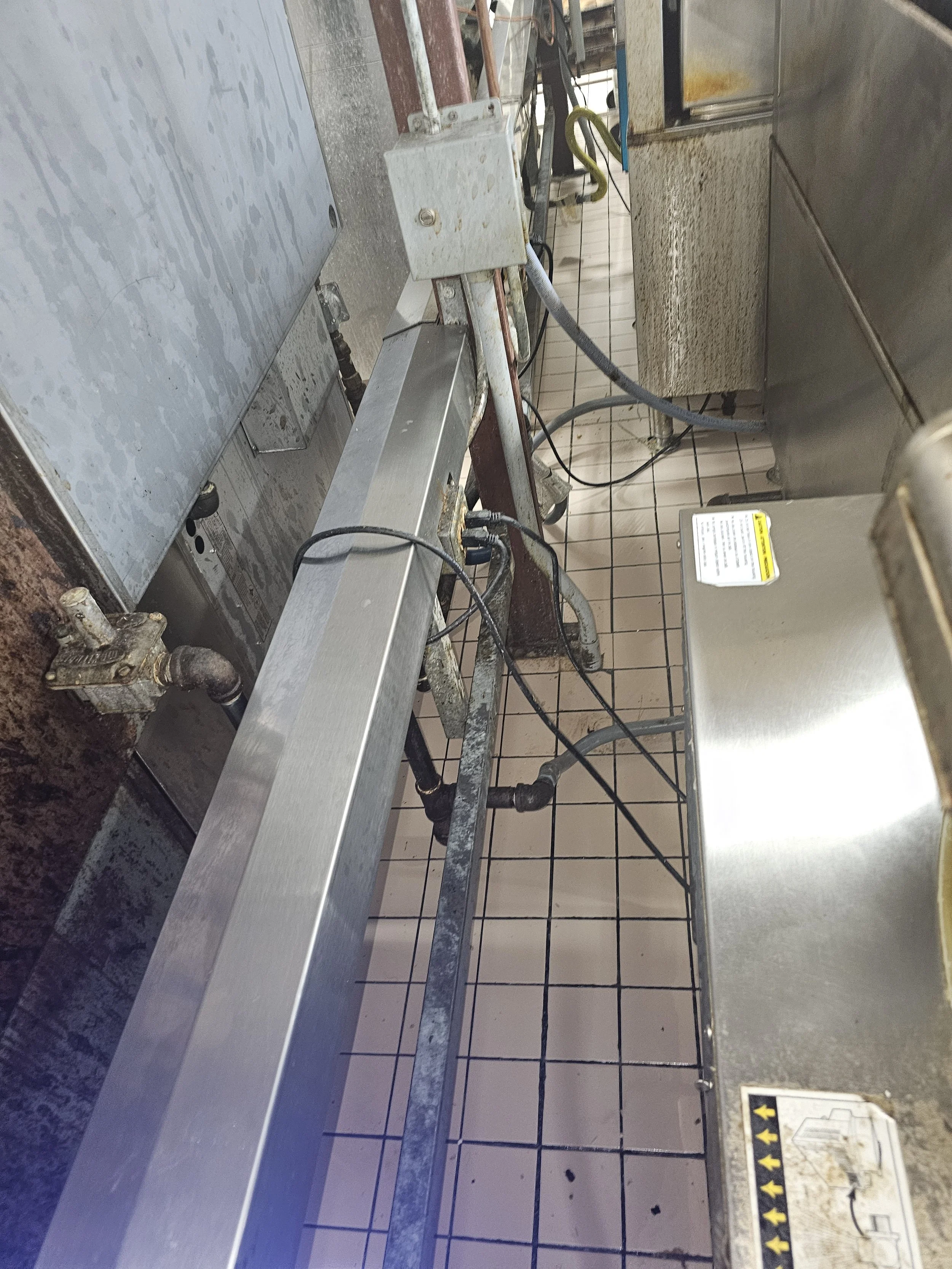 Commercial kitchen or industrial food prep area with stainless steel countertops, tiled floor, and exposed pipes and wiring.