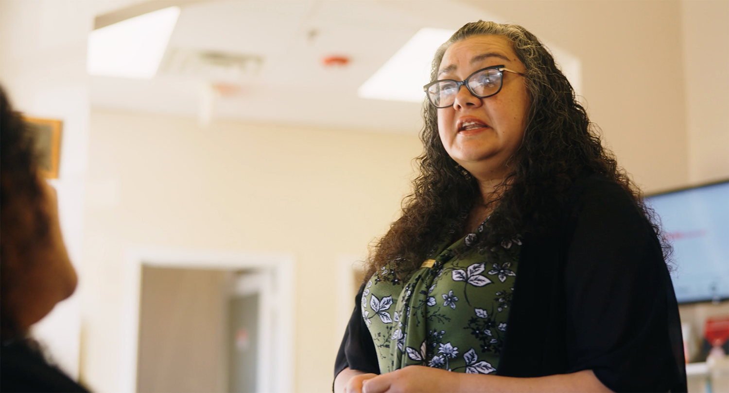 A woman with curly hair and glasses speaking to someone in an indoor setting, possibly an office or classroom.