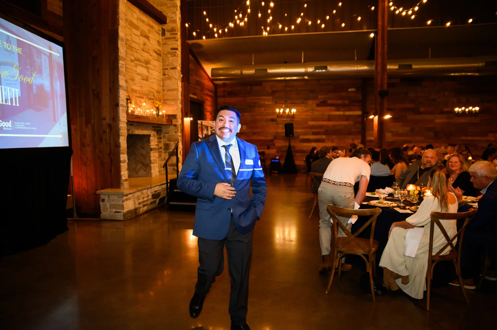 A man in a blue suit and tie stands smiling at an event in a rustic, warmly lit venue. The background shows a group of people seated at tables with place settings, a fireplace, and string lights.