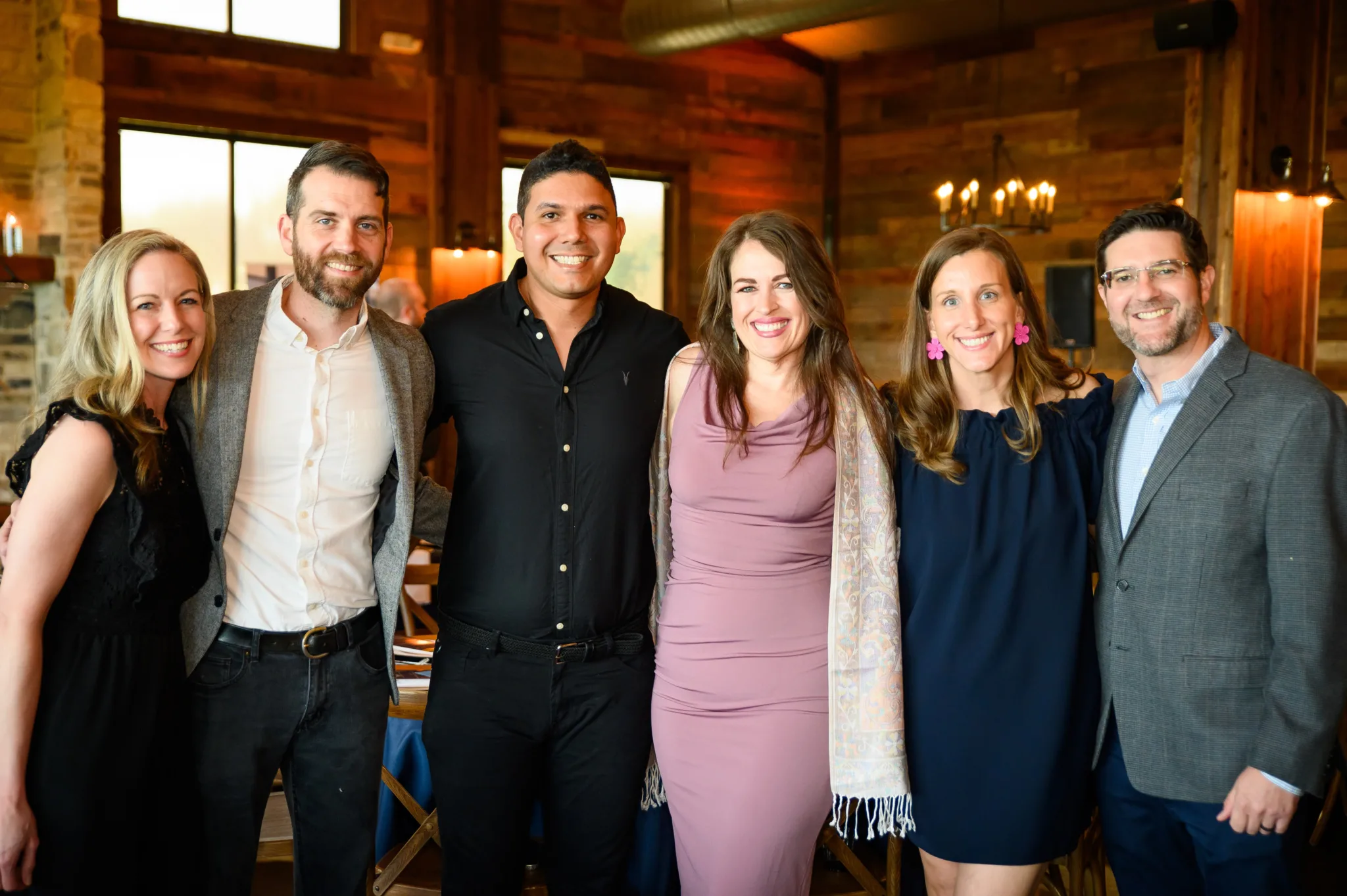 A group of six people smiling and standing close together in a warmly lit indoor setting, with wooden walls and large windows in the background.