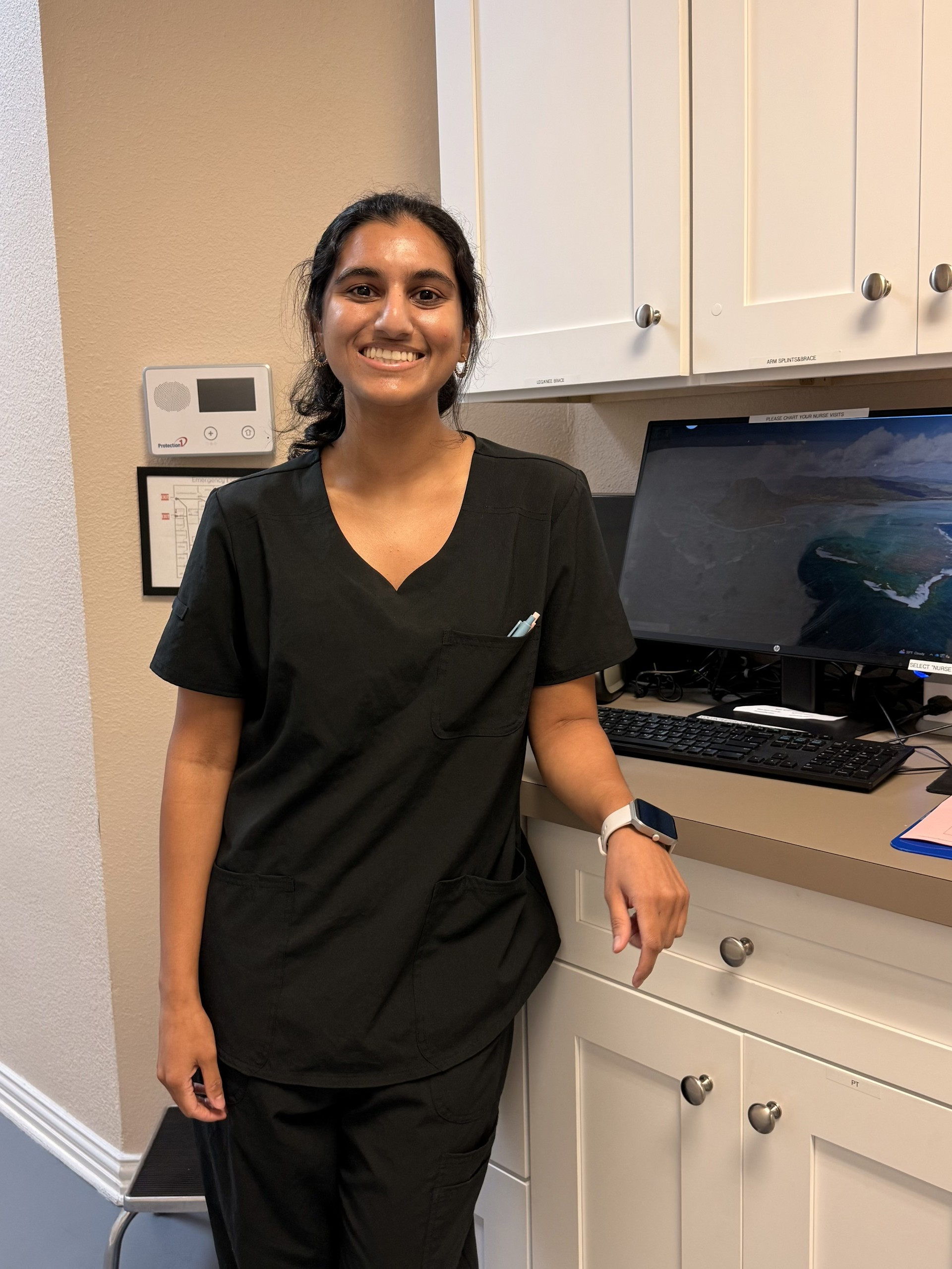 Smiling woman in black scrubs standing in a medical or office setting, leaning on a beige counter with a computer monitor, keyboard, and various office supplies.