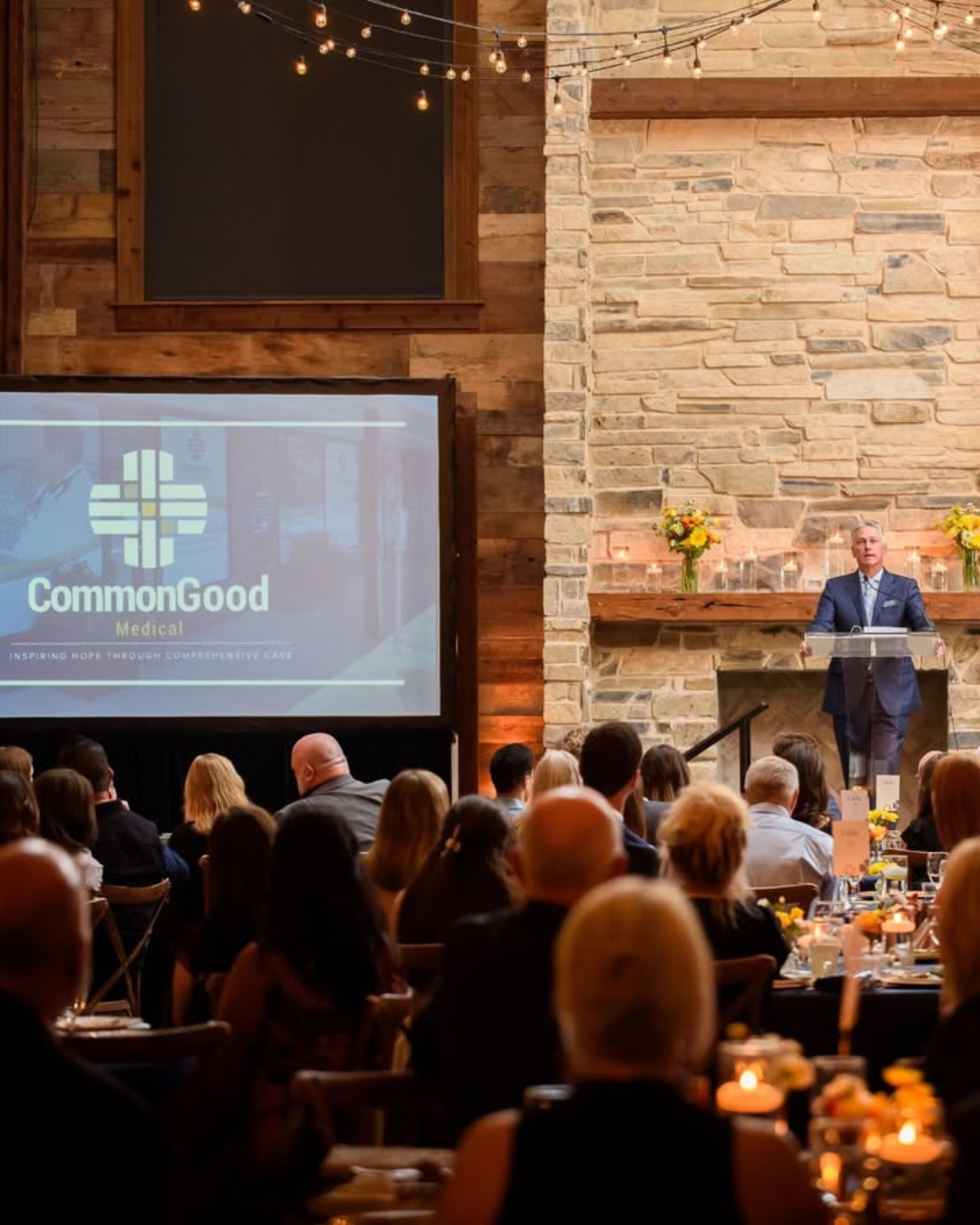A speaker in a suit delivers a speech at a podium decorated with flowers, at a event with an audience seated at tables, in a venue with a stone wall and string lights, for CommonGood Medical.