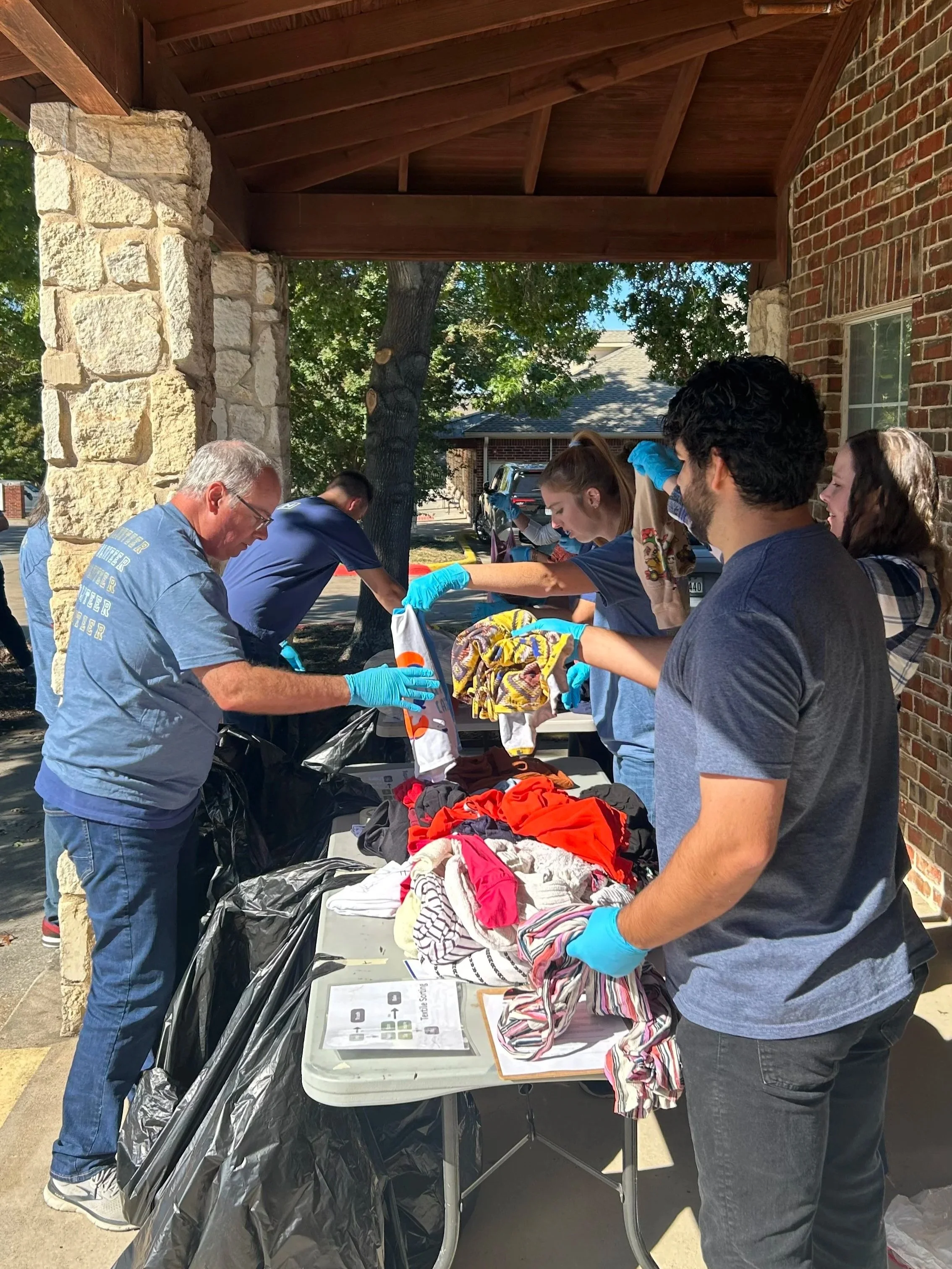 People sorting and bagging clothes at an outdoor charity event under a shelter with brick and stone columns.