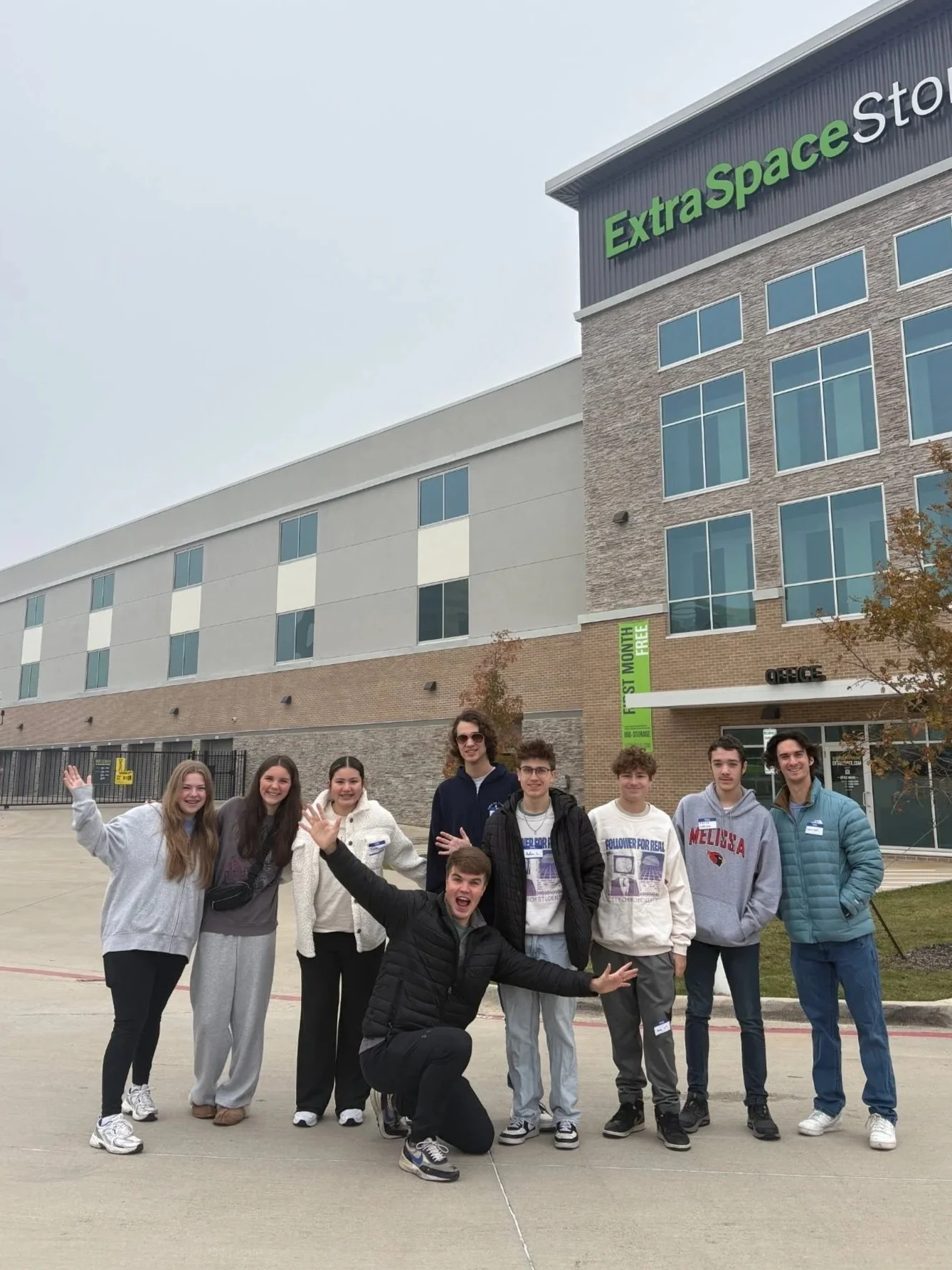 Group of young people posing in front of a large shopping complex called Extra Space Storage and Office, with some waving and others smiling, during the daytime.