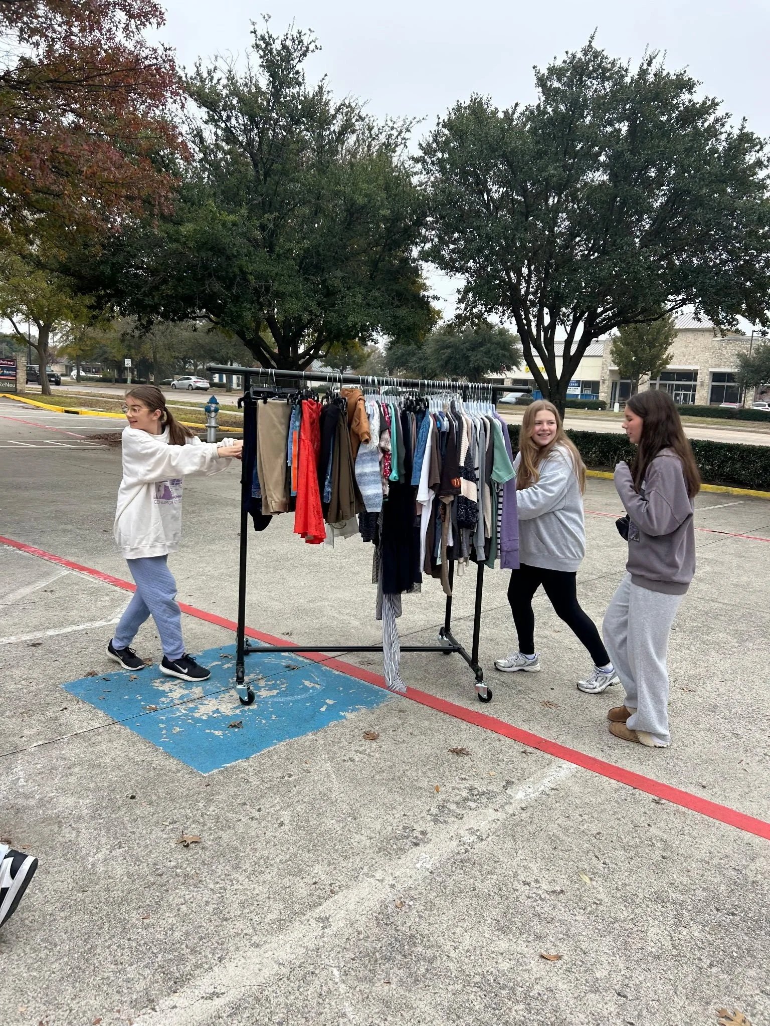 Three young women standing outdoors around a clothing rack with various clothes, talking and smiling, in a parking lot with trees and buildings in the background.