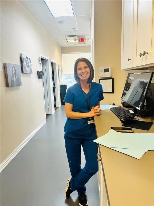 A smiling woman in blue medical scrubs standing at a reception desk in a healthcare facility.
