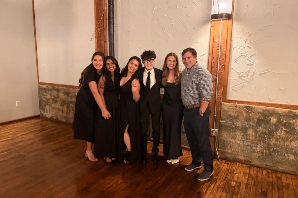 Group of six young adults smiling and posing together at a social event, with four women and two men dressed in semi-formal to formal attire, in an indoor setting with wooden floors and textured walls.