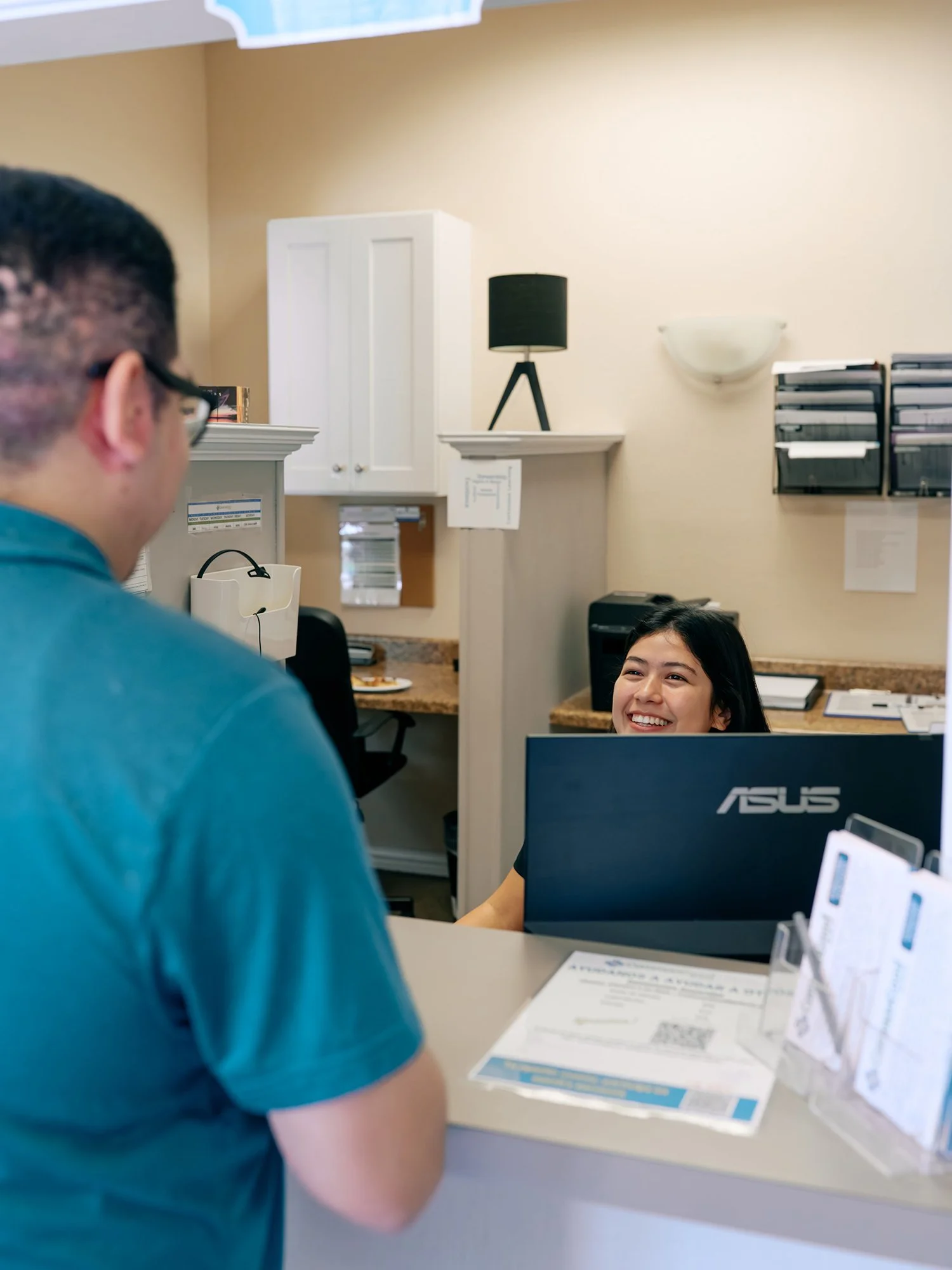 A man in a teal shirt at a reception desk speaking to a smiling female receptionist seated behind a computer with an ASUS logo. The background features file organizers, a lamp, and various office supplies.