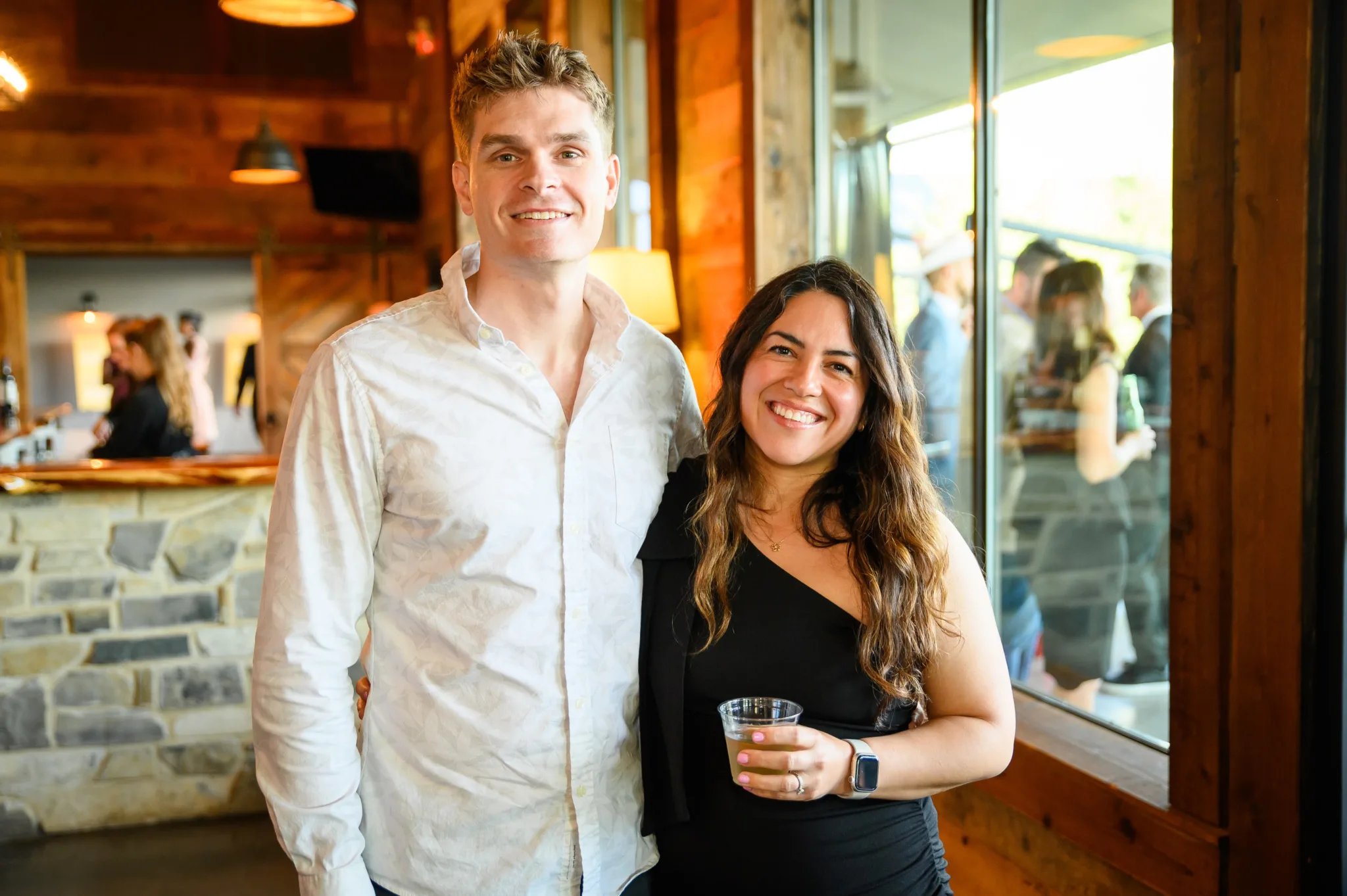 Smiling man in a white shirt and smiling woman in a black dress, holding a drink, standing inside a restaurant or bar with wooden decor and large windows.