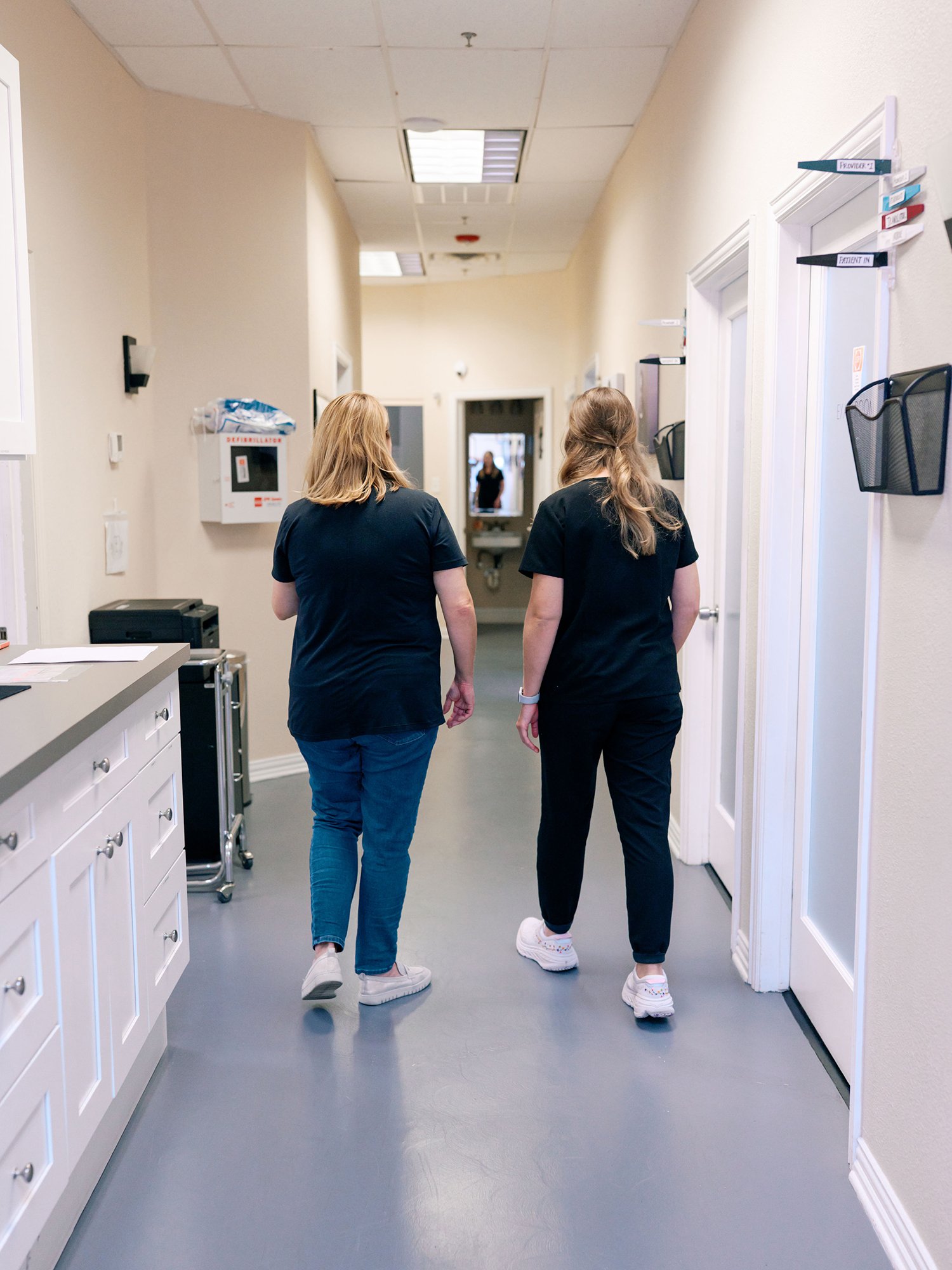 Two medical professionals walking down a corridor in a healthcare facility, seen from behind. They are dressed in dark scrubs, with one having blonde hair and the other with long, light brown hair. The corridor has white walls, closed doors with labels, and a gray floor.
