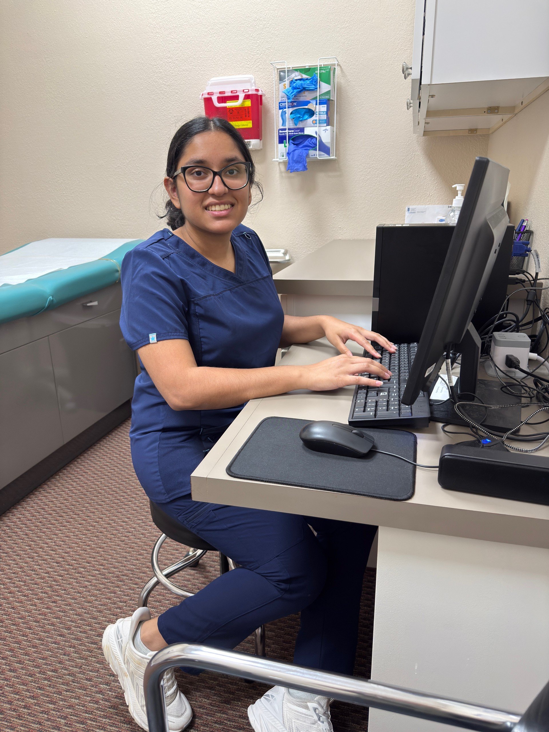 A woman with glasses wearing blue medical scrubs typing on a computer in a clinical office. She is smiling. There is a patient examination table with blue paper in the background and medical supplies on the wall.