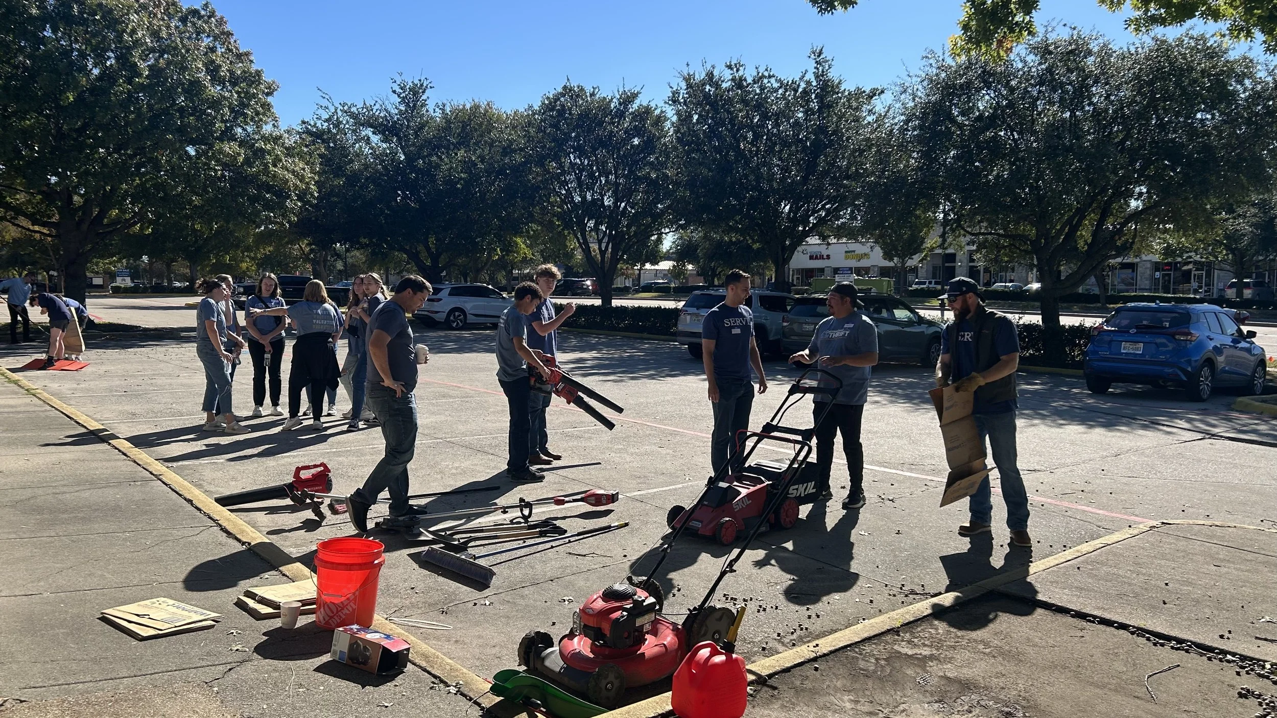 People working outdoors on a parking lot, surrounded by trees and parked cars, with tools and equipment scattered on the ground.