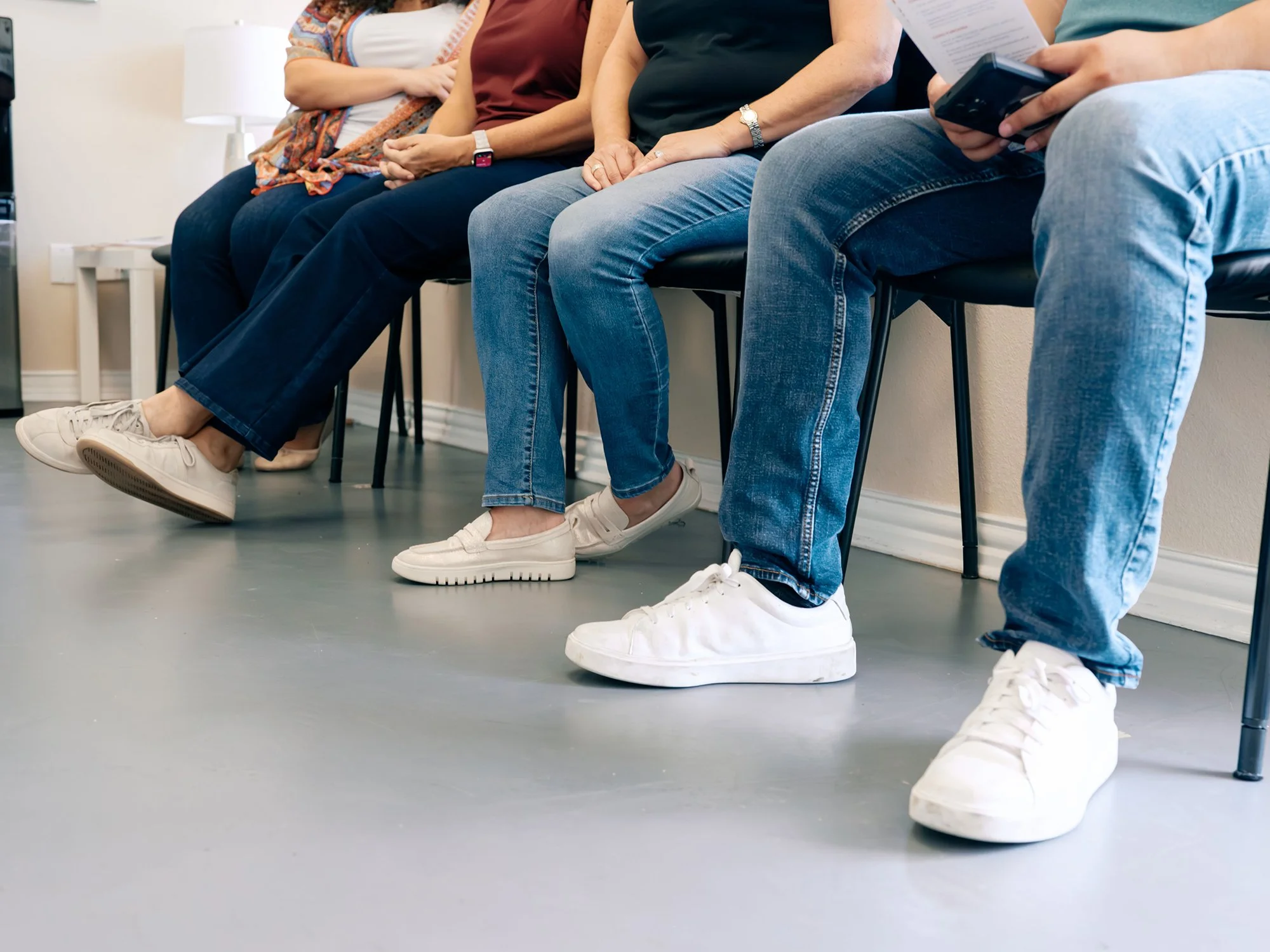 People sitting on chairs waiting, wearing casual clothes and white sneakers, with some holding phones or papers, in a room with a gray floor and white walls.
