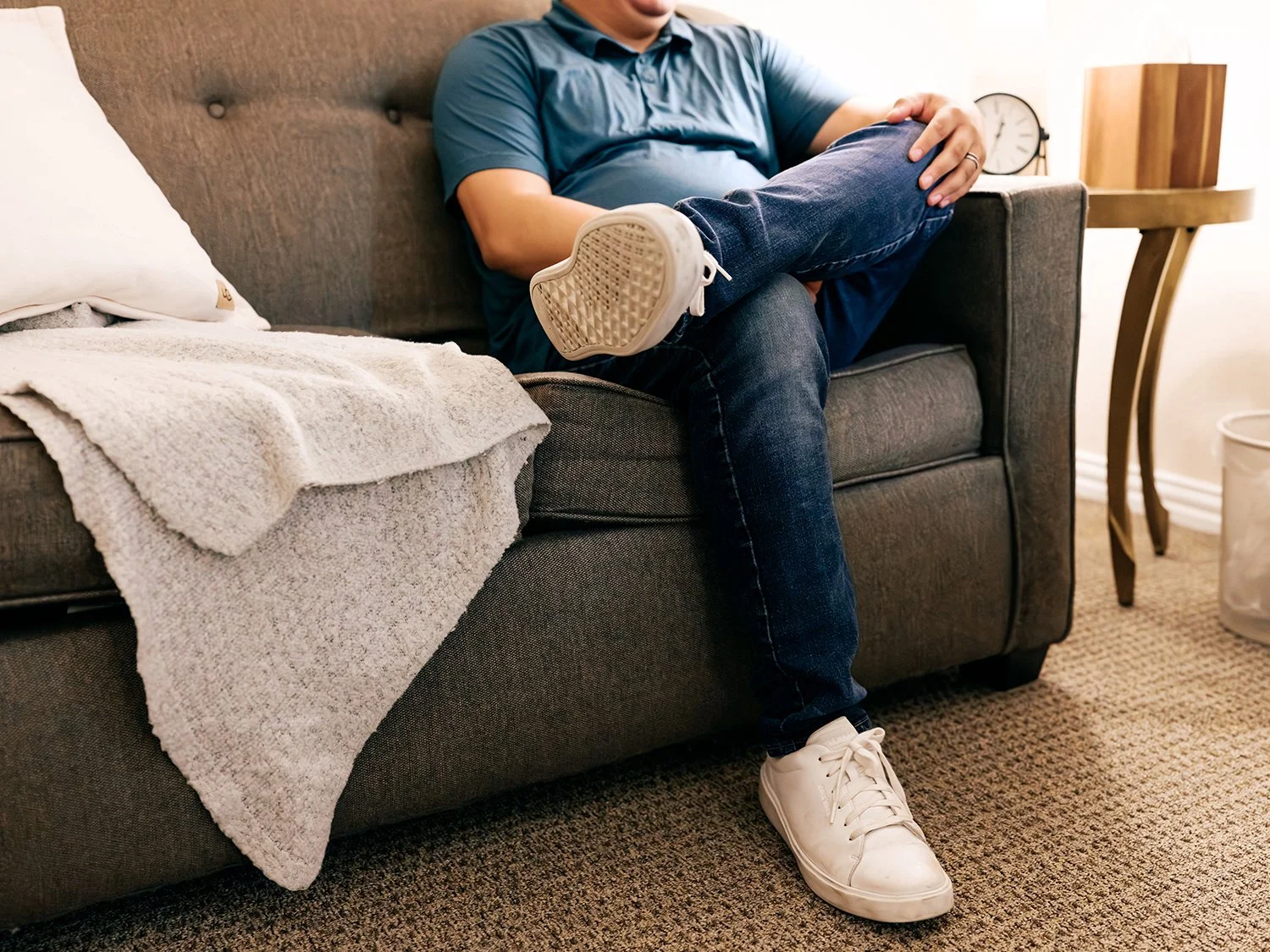 A person sitting on a gray sofa with one leg crossed over the other, wearing a blue shirt, jeans, and white sneakers. There is a beige throw blanket draped over the arm of the sofa, and a white pillow behind the person. A wooden side table with a clock and books is in the background, along with a beige carpeted floor.