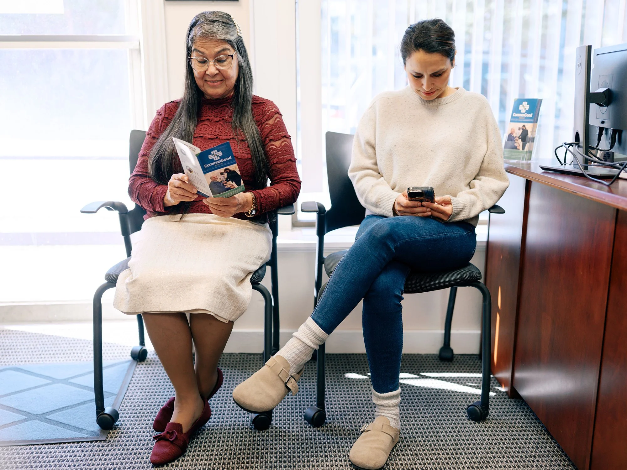Two women sitting in chairs in a waiting room, looking at their phones. The woman on the left has long gray hair, glasses, and is wearing a red lace top and a beige skirt. The woman on the right has dark hair pulled back, wearing a beige sweater, blue jeans, and beige slippers.