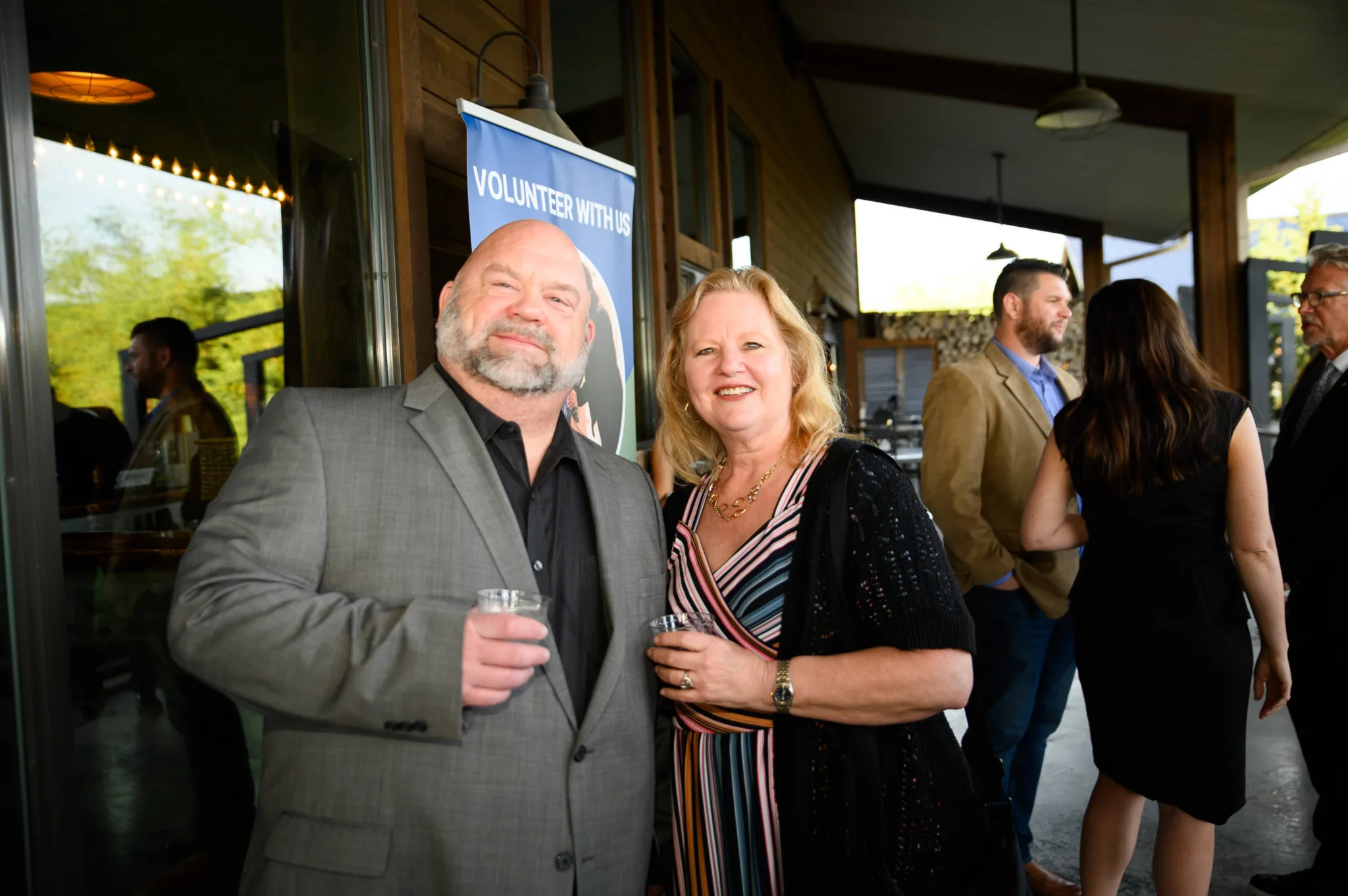 People attending a social event outdoors, with a man and a woman in the foreground smiling and holding drinks, a poster behind them encouraging volunteering, and others socializing in the background.