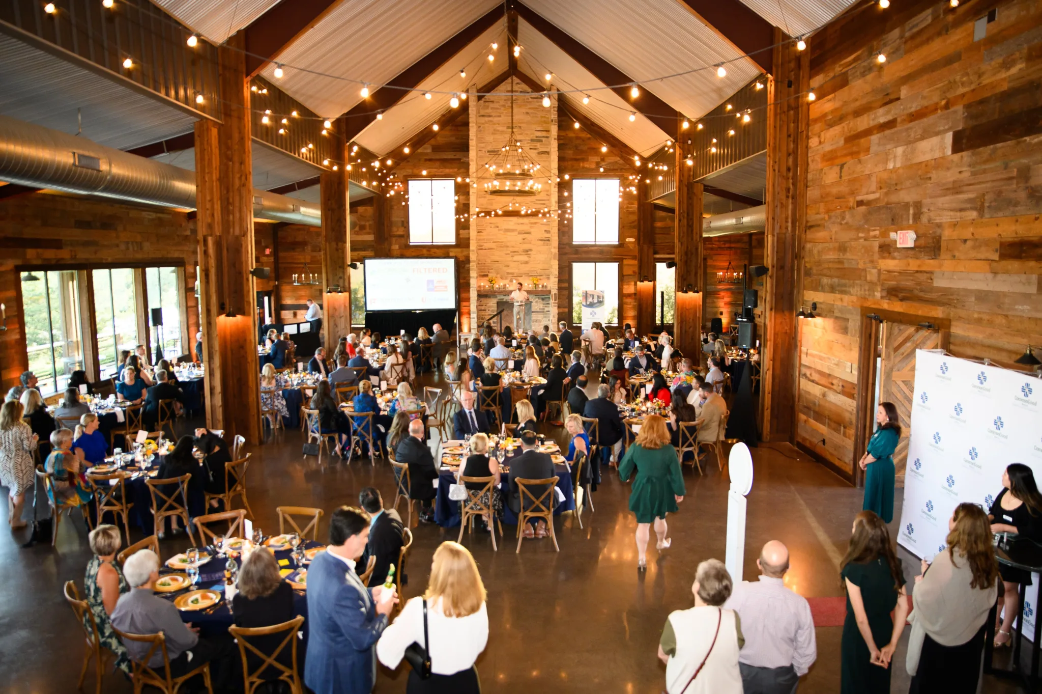 A large indoor gathering with many people seated at round tables in a rustic wooden hall with string lights overhead and large windows in the background. A woman stands at a podium in front of a fireplace, delivering a presentation, and there are mul