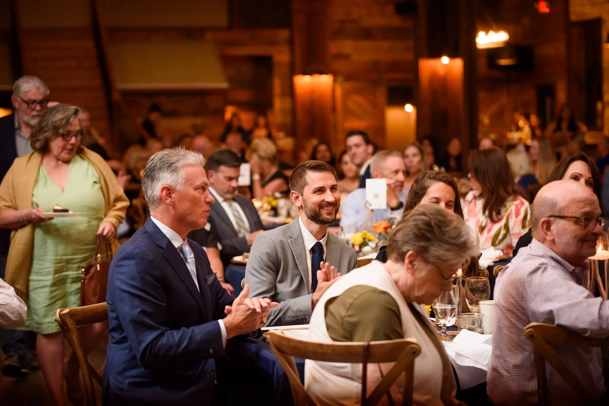 People attending a formal event or dinner in a warm, wood-paneled room, seated at tables with floral centerpieces, some engaged in conversation, others listening, with a woman serving food in the background.