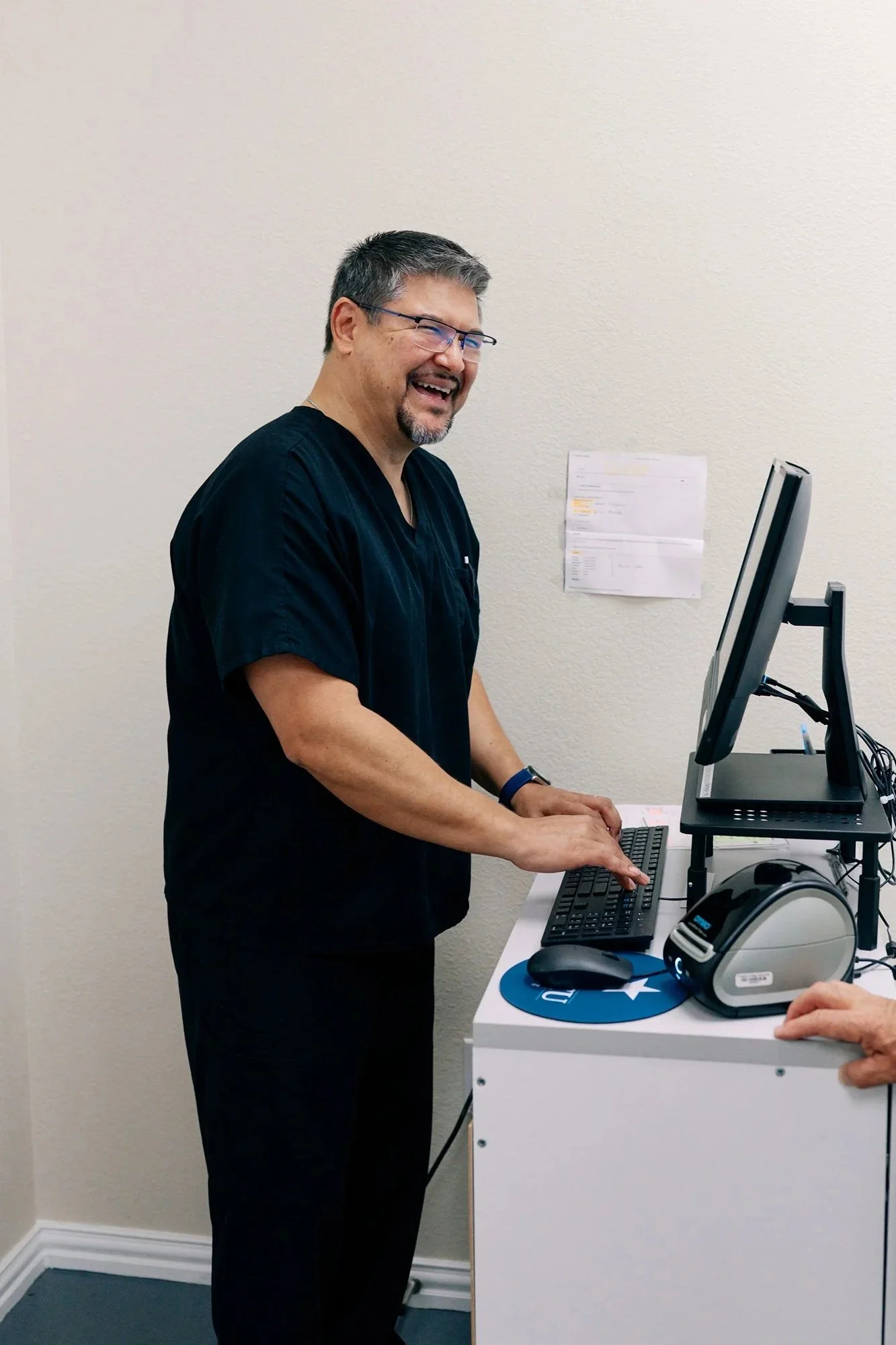A man with glasses, a beard, and gray hair wearing black scrubs, smiling while working on a computer in an office with a light-colored wall and a paper posted on it.