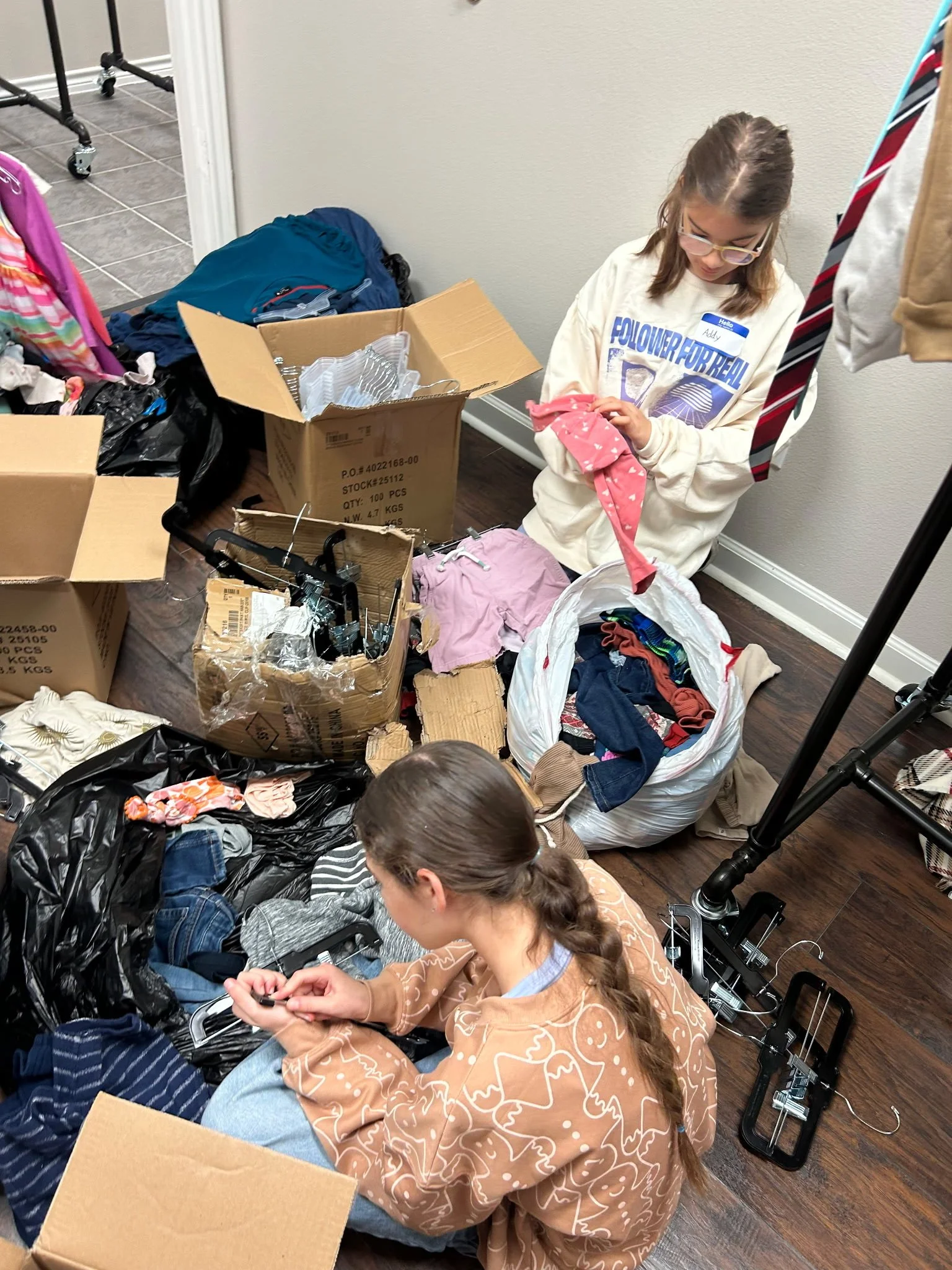 Two girls and a boy sorting through clothes and hangers in a room filled with boxes and bags, some clothing, and a clothing rack.