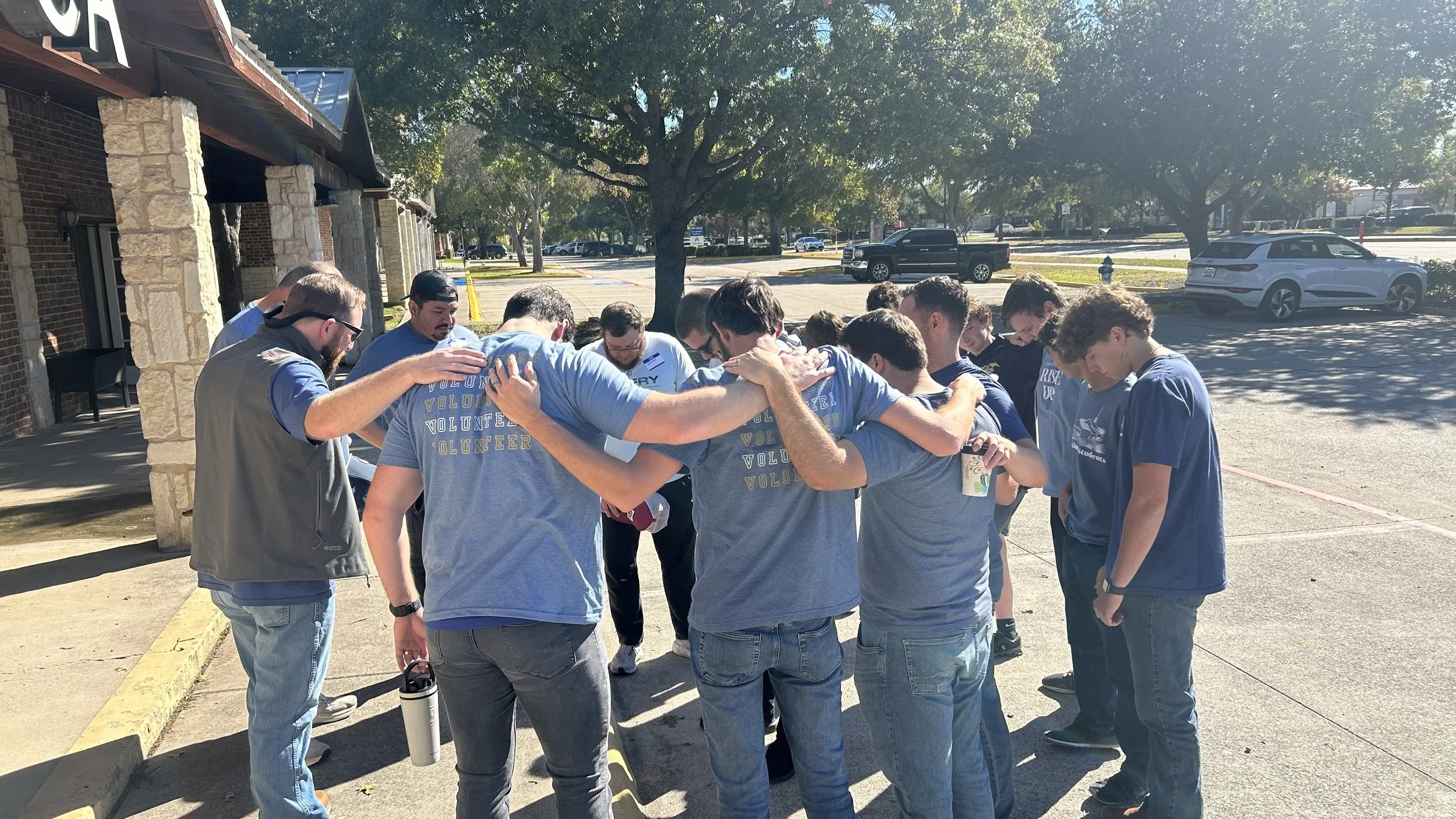 Group of people standing in a circle outdoors with heads bowed, praying or reflecting, in a parking lot next to a building and trees.