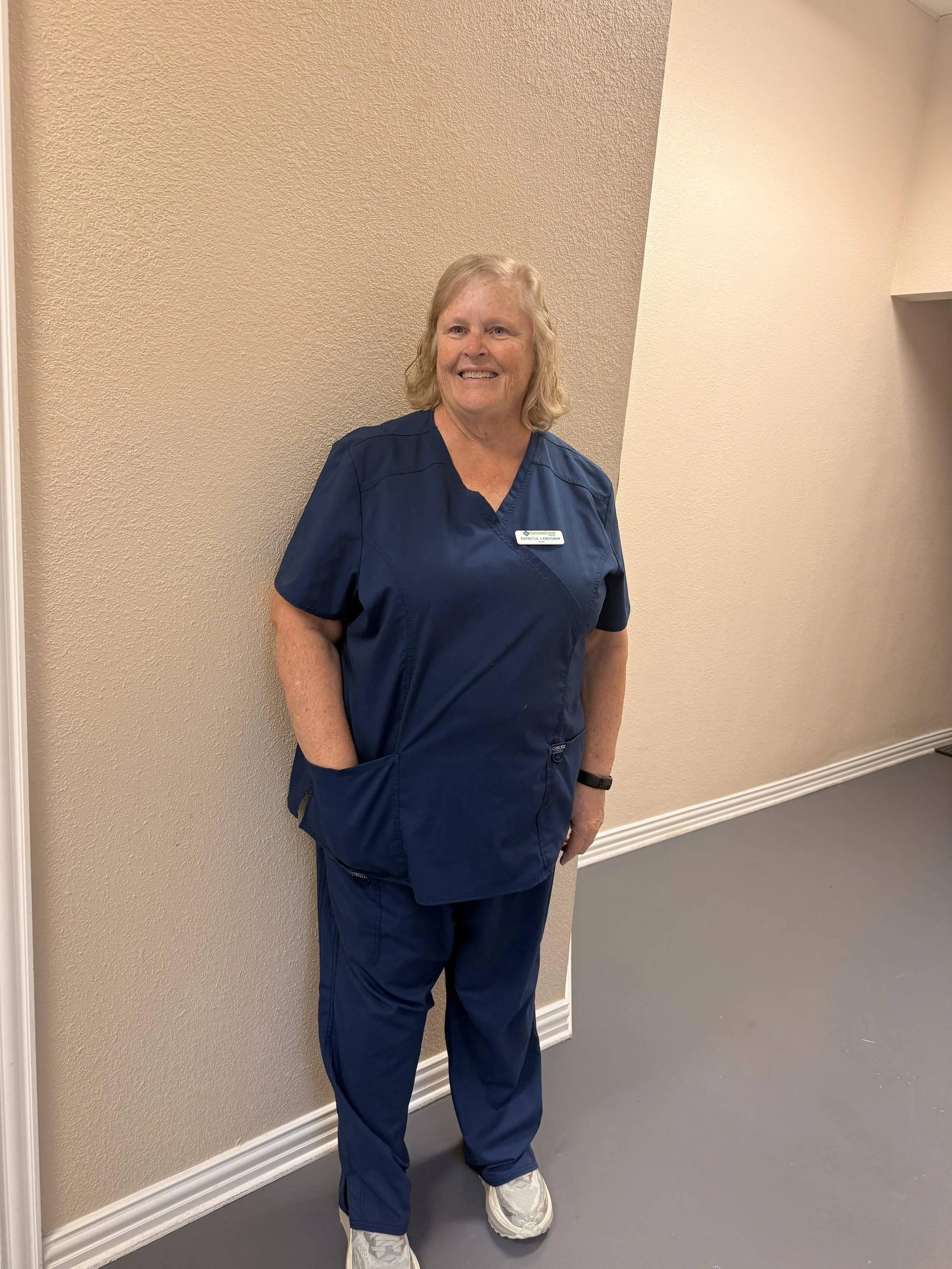 A smiling woman with blond hair wearing navy scrubs and white sneakers standing against a beige textured wall with a white baseboard.