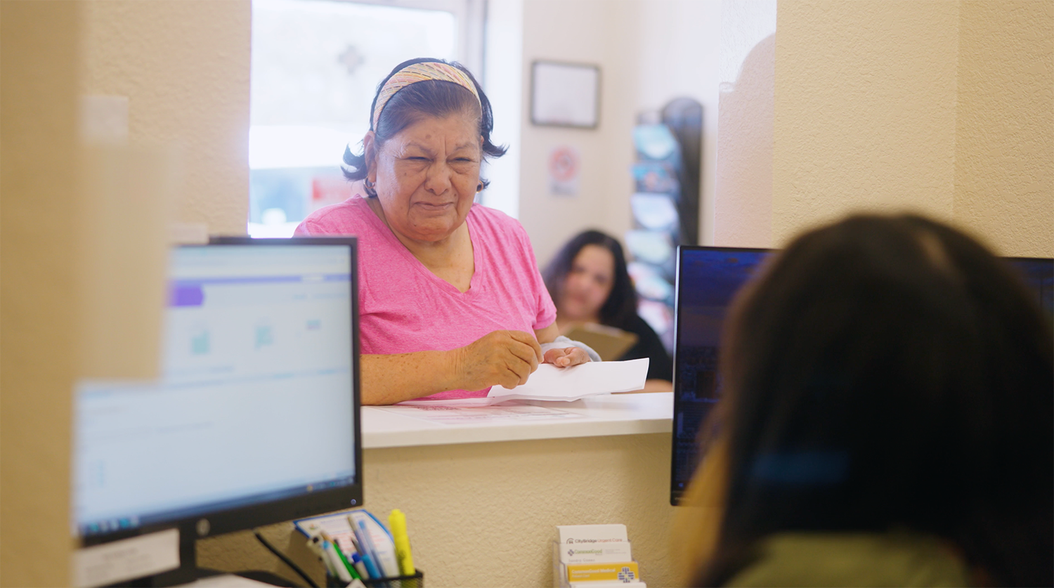 An elderly woman at a reception window holding papers, talking to a woman with long dark hair seated at a computer.