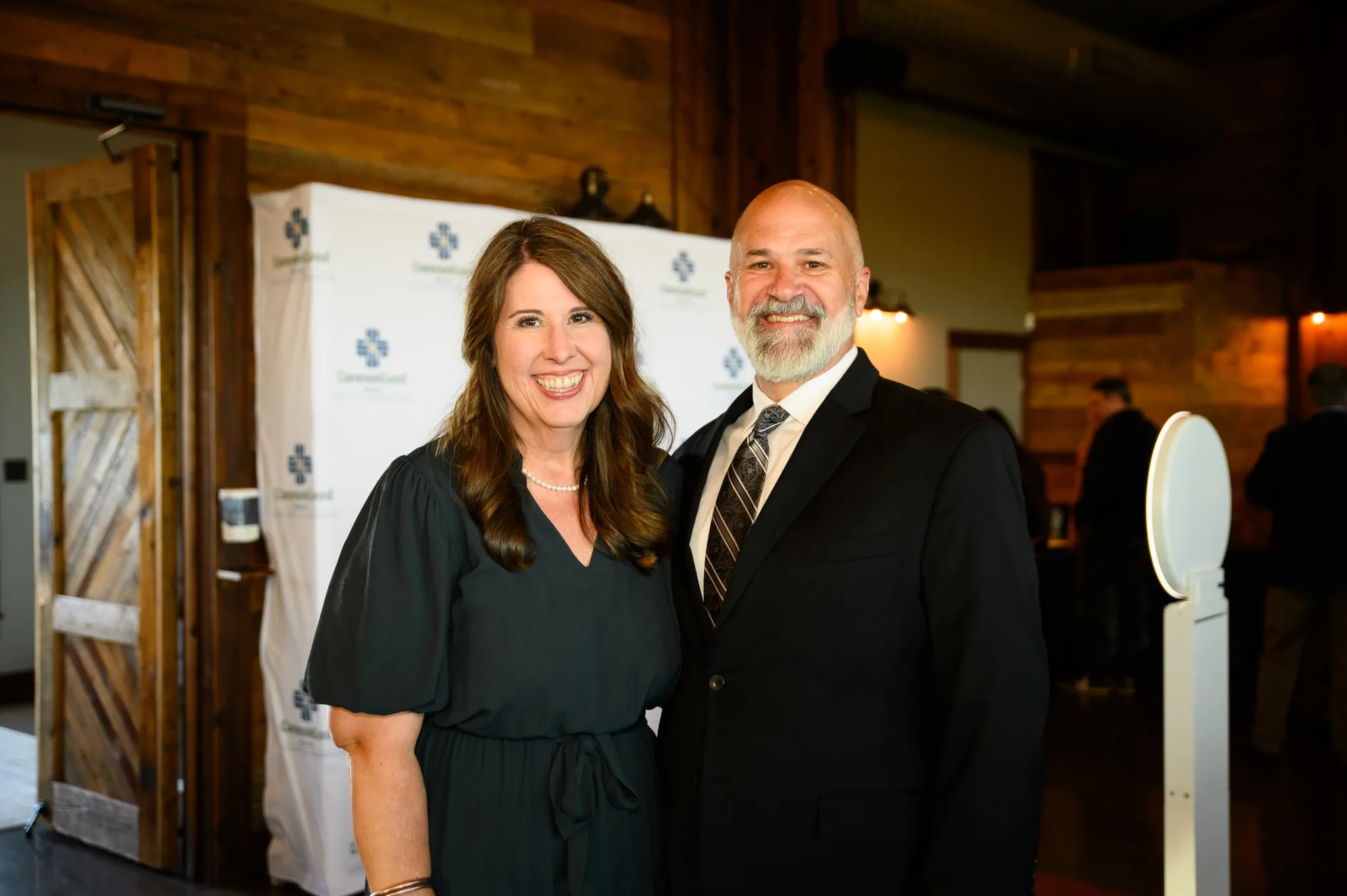 A woman and a man dressed in formal attire, smiling at the camera at an indoor event with wooden walls and a branded backdrop.