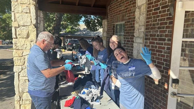 A group of people volunteering at a clothes drive, sorting and organizing clothes on a table outside a brick building. Everyone is wearing gloves, and some are smiling or laughing.
