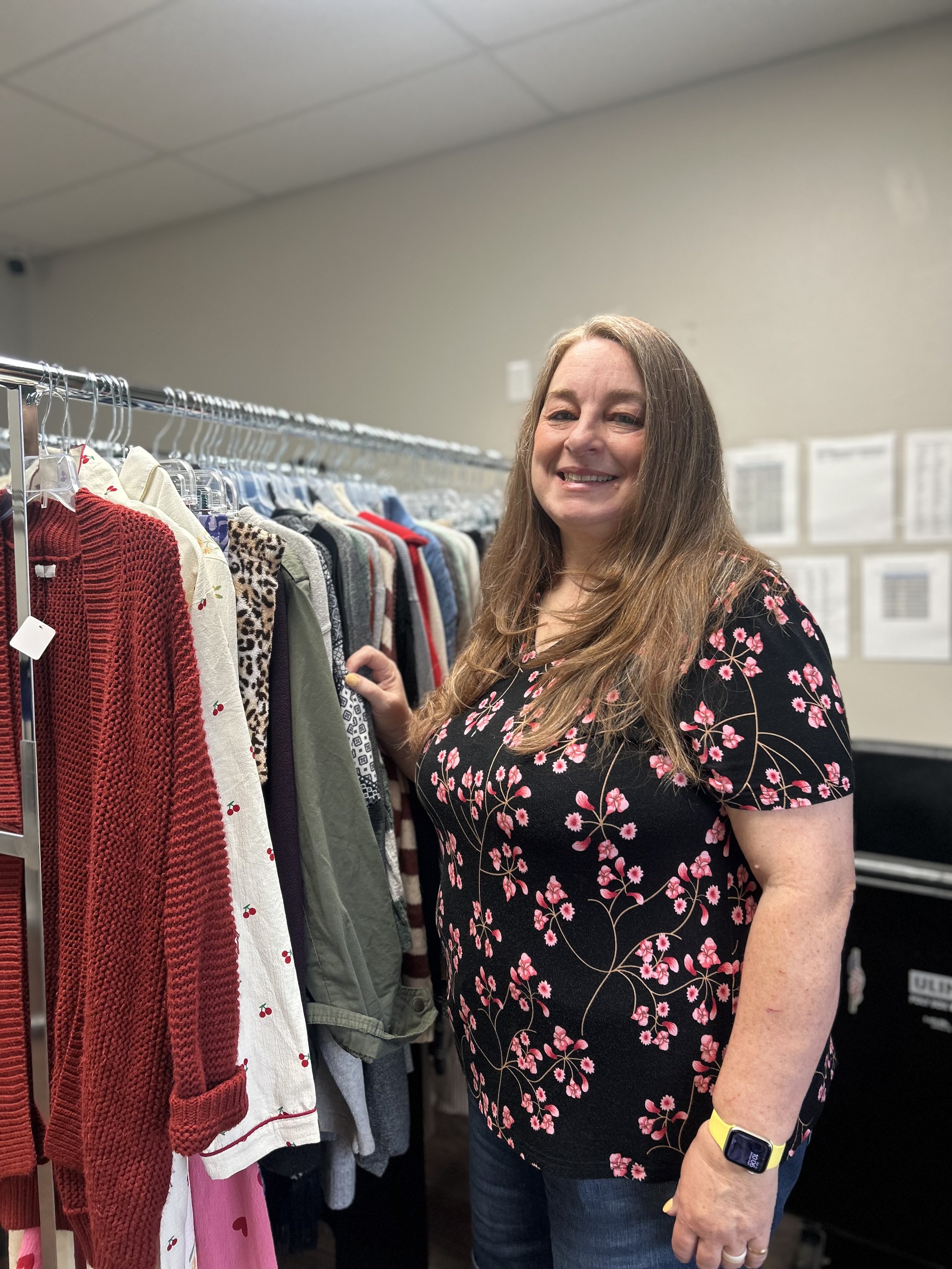 A woman with long brown hair wearing a black dress with pink floral designs is shopping in a clothing store and looking at sweaters on a rack.