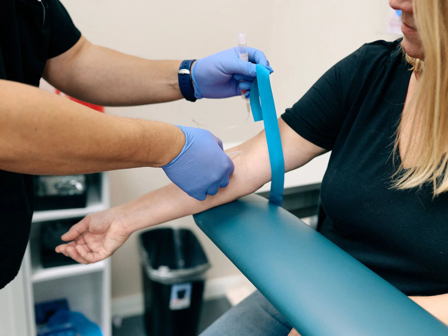 Person in black shirt receiving blood draw from healthcare professional wearing gloves, using a needle and vials in a clinical setting.