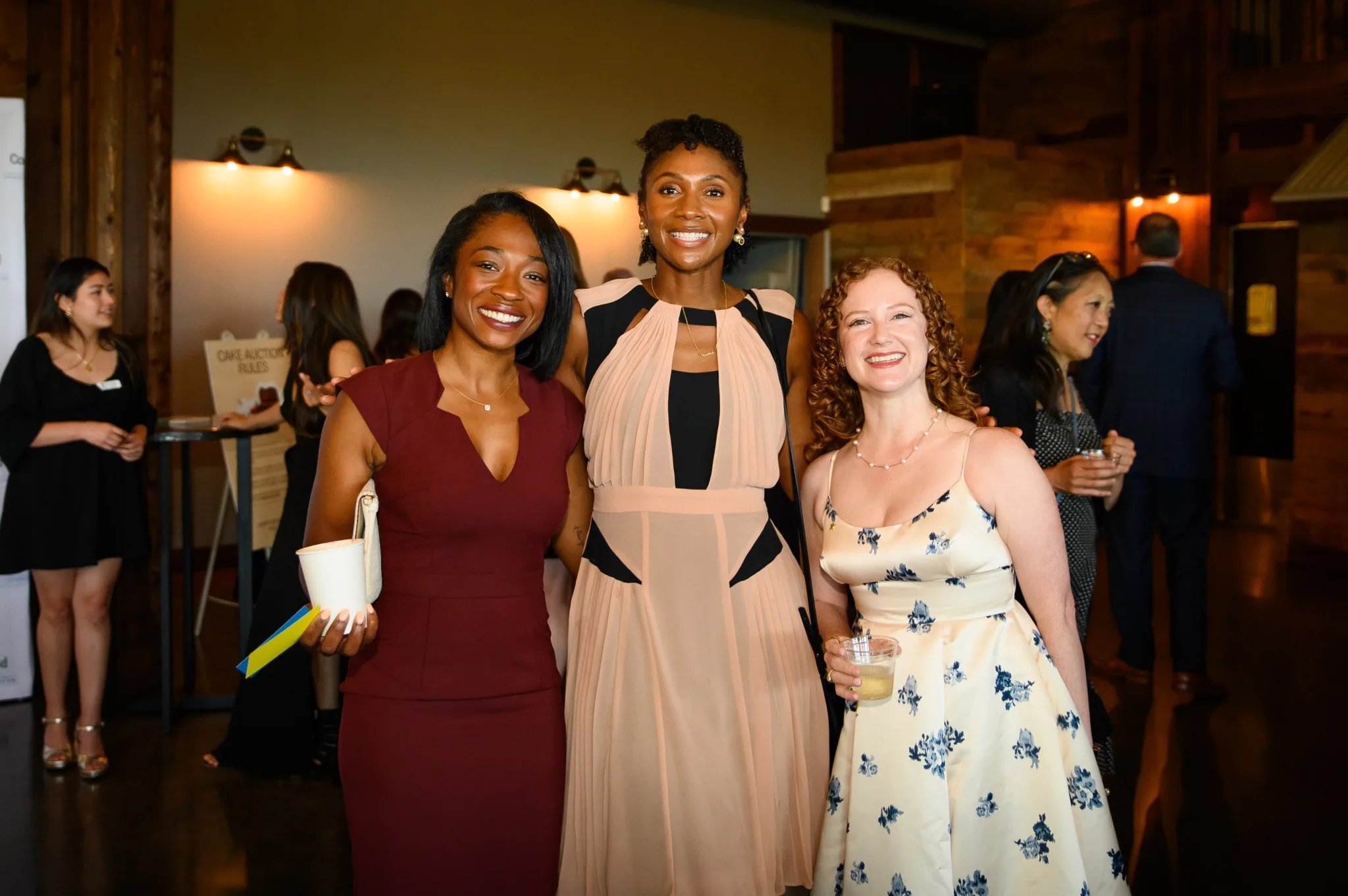 Three women smiling and posing together at an indoor event. The woman on the left is wearing a burgundy dress, holding a drink and papers. The woman in the middle is wearing a multicolored striped dress with earrings. The woman on the right is wearin